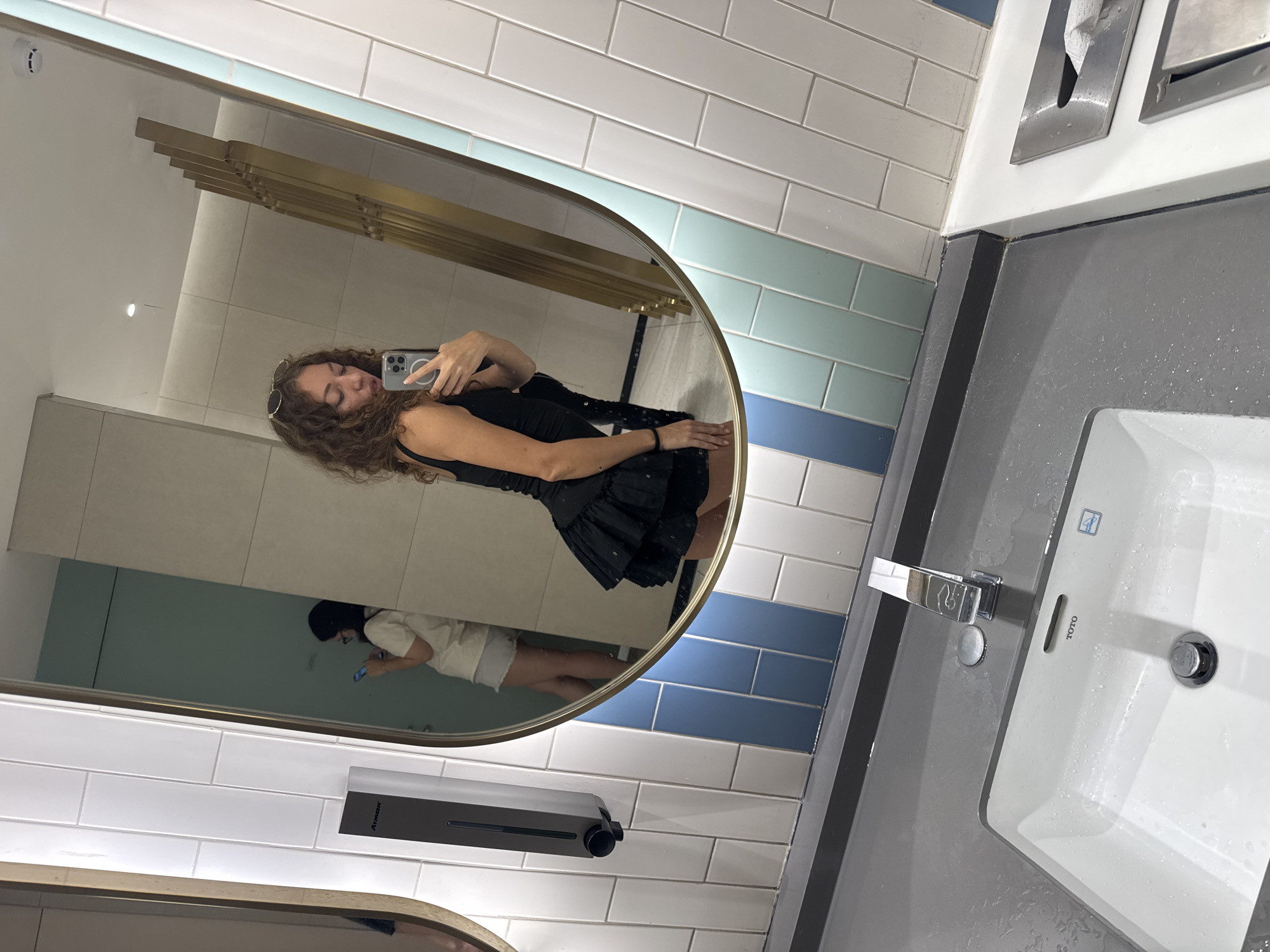 A woman is taking a selfie in a modern public restroom, likely in Hanoi, Vietnam, based on the context. She is wearing a black dress and sunglasses pushed up on her head, and is holding a smartphone. In the mirror's reflection, another woman is visible in the background, also looking at her phone. The restroom features a white sink, a chrome faucet, and a grey countertop. The walls are tiled with a mix of white, blue, and light green rectangular tiles, and there is a soap dispenser mounted on the wall. The lighting is bright and even, suggesting an indoor environment.