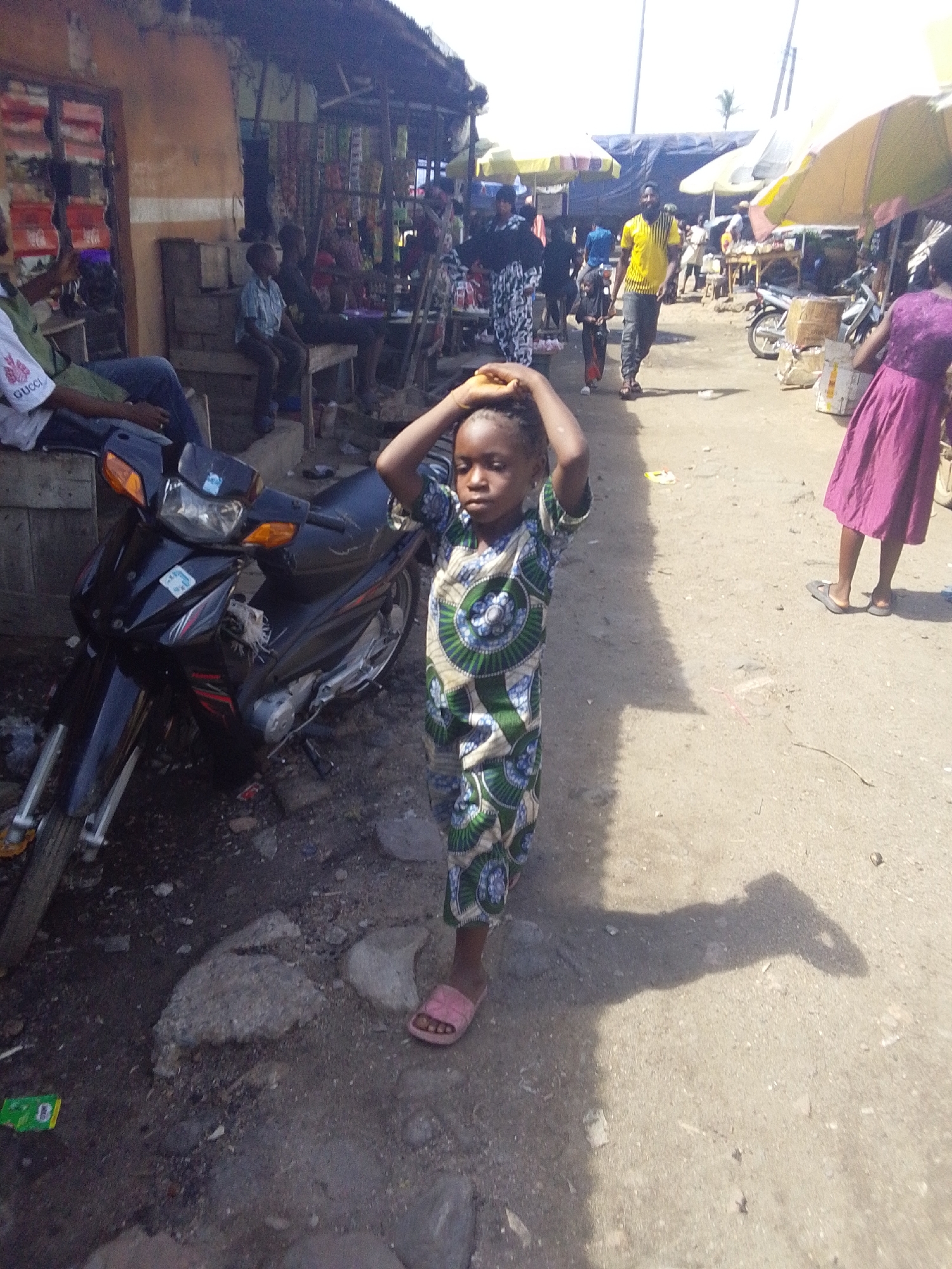 The image presents a vibrant outdoor market scene in Lokoja, Nigeria, captured under bright daylight which casts strong shadows on the dusty ground, suggesting midday.
The central subject is a young child, possibly female, standing with her hands clasped on top of her head. She wears a striking, patterned one-piece outfit in shades of green, blue, and white, adorned with circular and wave motifs, and light pink slip-on sandals. Her expression is neutral as she looks towards the viewer.
To the child's immediate left, a black motorcycle is parked. Further left, against a building with a reddish-brown facade, several individuals are seated on wooden benches, obscured by market structures. One person in the foreground wears a white t-shirt with the word "GUCCI" visible on the sleeve. Behind them, market stalls, possibly made of wood and corrugated metal, display various items for sale.
To the child's right, the market continues, populated by other people moving about. Several large yellow and white market umbrellas provide shade over different stalls. A person in a long purple dress is visible from the back on the far right. In the middle distance, a man in a bright yellow and black striped shirt walks towards the left side of the frame, past other market-goers and stalls.
The ground is unpaved, composed of dirt, small rocks, and scattered debris. The overall scene depicts a bustling, typical day in an informal market environment.