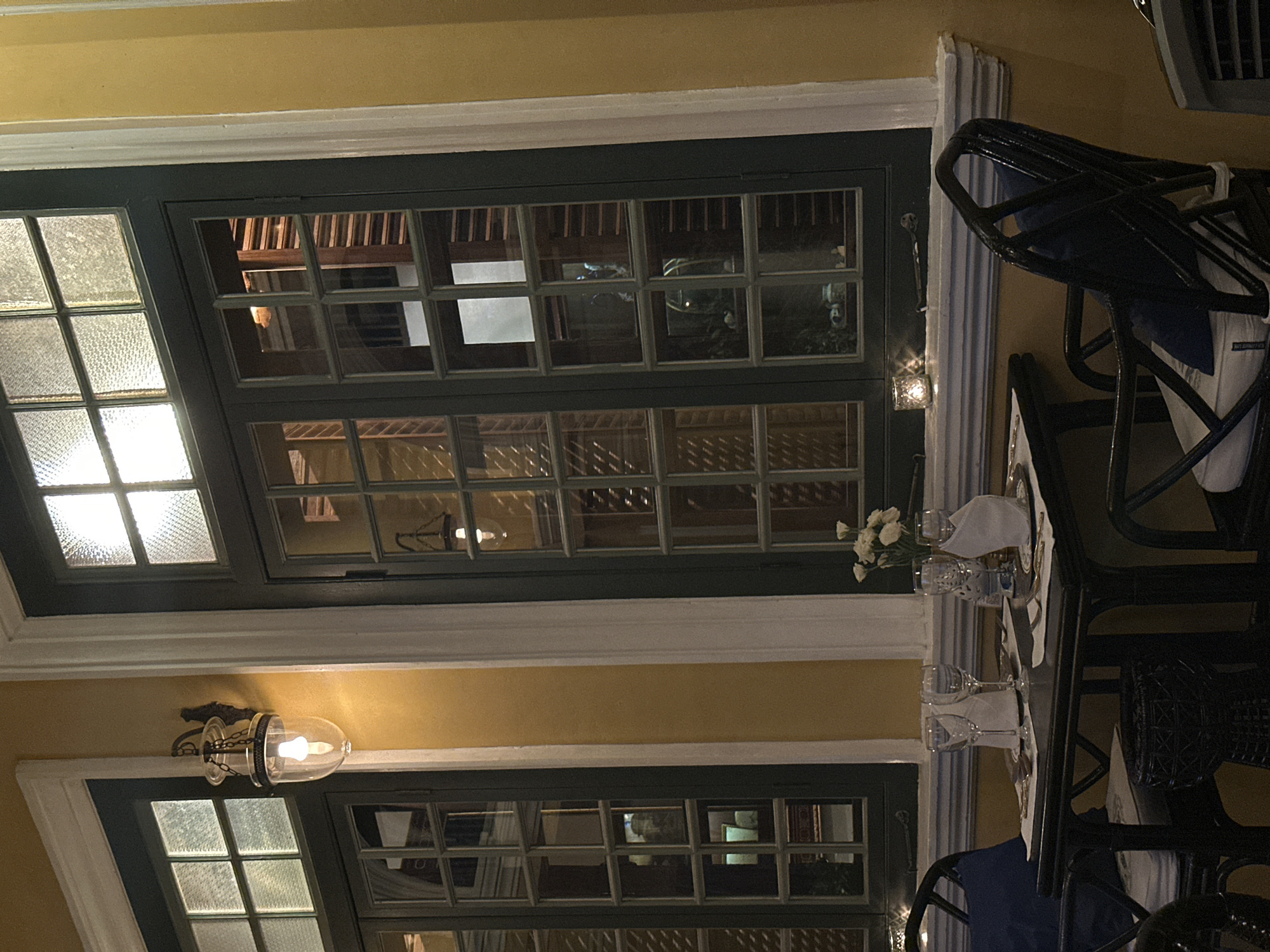 The image shows an interior view in Phuket City Municipality, Thailand, featuring a dining table set with glasses and a floral arrangement, alongside chairs, positioned near a large window with multiple panes. The lighting appears dim, highlighting the details of the table setting and the window's architectural design. There are no people visible in the immediate scene. A wall sconce is also visible.