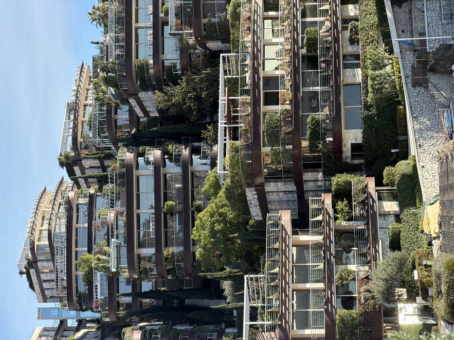 The image displays a complex of multi-story, terraced buildings situated on a steep, verdant hillside in Boreti, Montenegro. The structures feature a contemporary architectural design with curvilinear forms, characterized by extensive glass window walls, light-colored stone cladding, and dark brown facade elements, all bordered by silver-toned metal railings on numerous balconies and terraces. Abundant greenery, including tall, slender evergreen trees, various shrubs, and potted plants, is integrated throughout the property and on individual terraces, some of which are covered by white metal pergolas. Several balconies contain outdoor furniture such as chairs, tables, and sun loungers. An external glass elevator shaft is visible on the left side of one building. In the lower foreground, a stone retaining wall is prominent, below which an area of disturbed earth is partially covered by a yellow tarpaulin or netting. Two green waste bins are positioned near this area. Further down, a small, light blue boat rests on or near a lower section of the stone wall, adjacent to steps leading downwards. A flagpole with a partially visible blue flag stands near the water's edge. The scene is illuminated by clear daylight, and no individuals are visible within the frame.