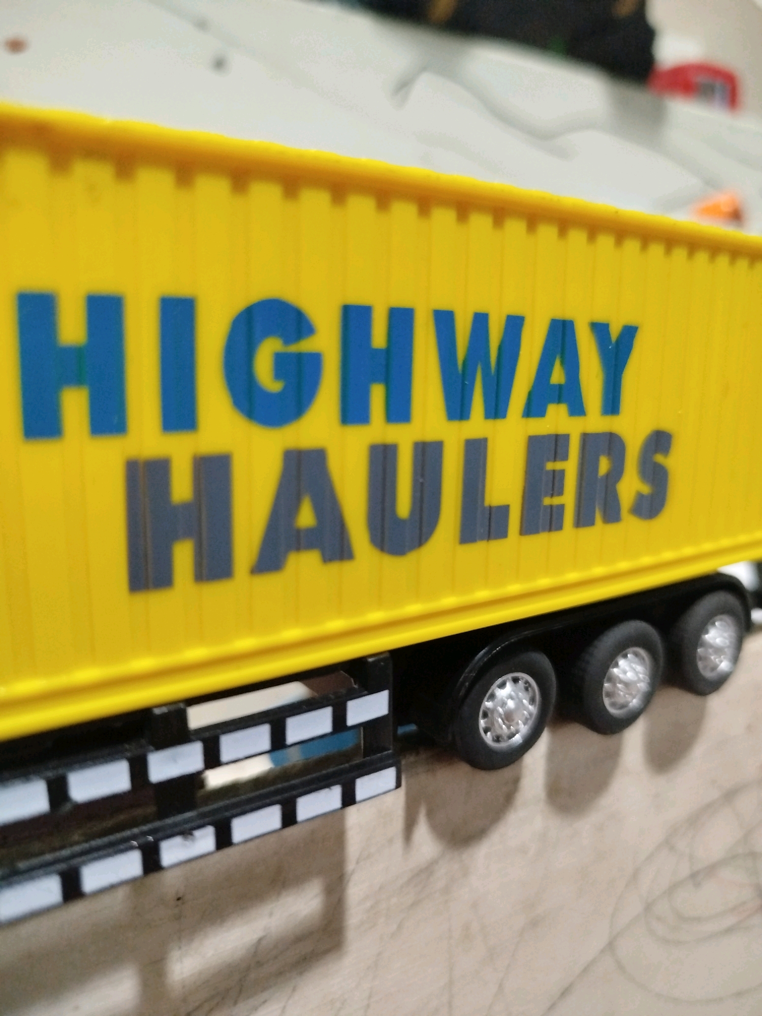The image displays a close-up, angled shot of a yellow toy truck trailer. The trailer has "HIGHWAY HAULERS" printed on its side in large blue and gray lettering. The wheels of the trailer are visible at the bottom of the image, showing a row of three silver-rimmed black tires. The trailer appears to be on a light-colored, possibly wooden surface. The background is blurred, but suggests an indoor environment with some white and dark elements, possibly indicating other toys or objects. There are no people or specific landmarks visible in the image, therefore its location in Mojoagung, Indonesia cannot be determined from the visual information provided.