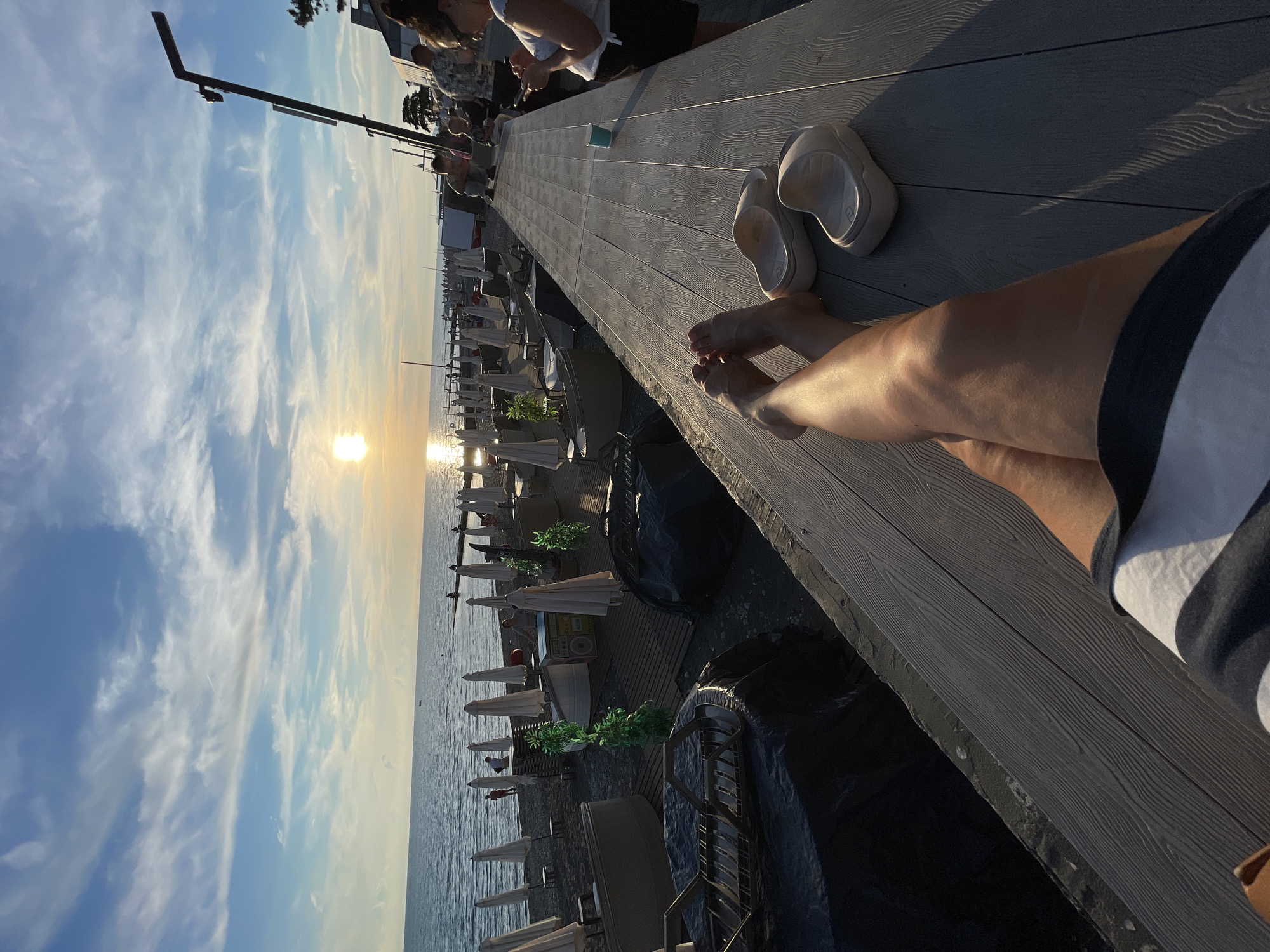 This image depicts a sunset at a beachside establishment in Sochi, Russia. The foreground features a person's legs and feet resting on a wooden deck, with a pair of light-colored slides nearby. The person appears to be wearing striped shorts. The midground shows a beach lined with white umbrellas and lounge chairs, indicating a public beach area. Several people are visible in the distance, some walking along the shore and others seated. Beyond the beach, the Black Sea stretches to the horizon, with the sun setting, casting a warm glow. The sky is a mix of blue and white clouds, with the sun prominently visible. To the right, a modern lamppost stands tall, and part of a building or structure is visible. The overall atmosphere is one of relaxation and enjoyment of a seaside evening.