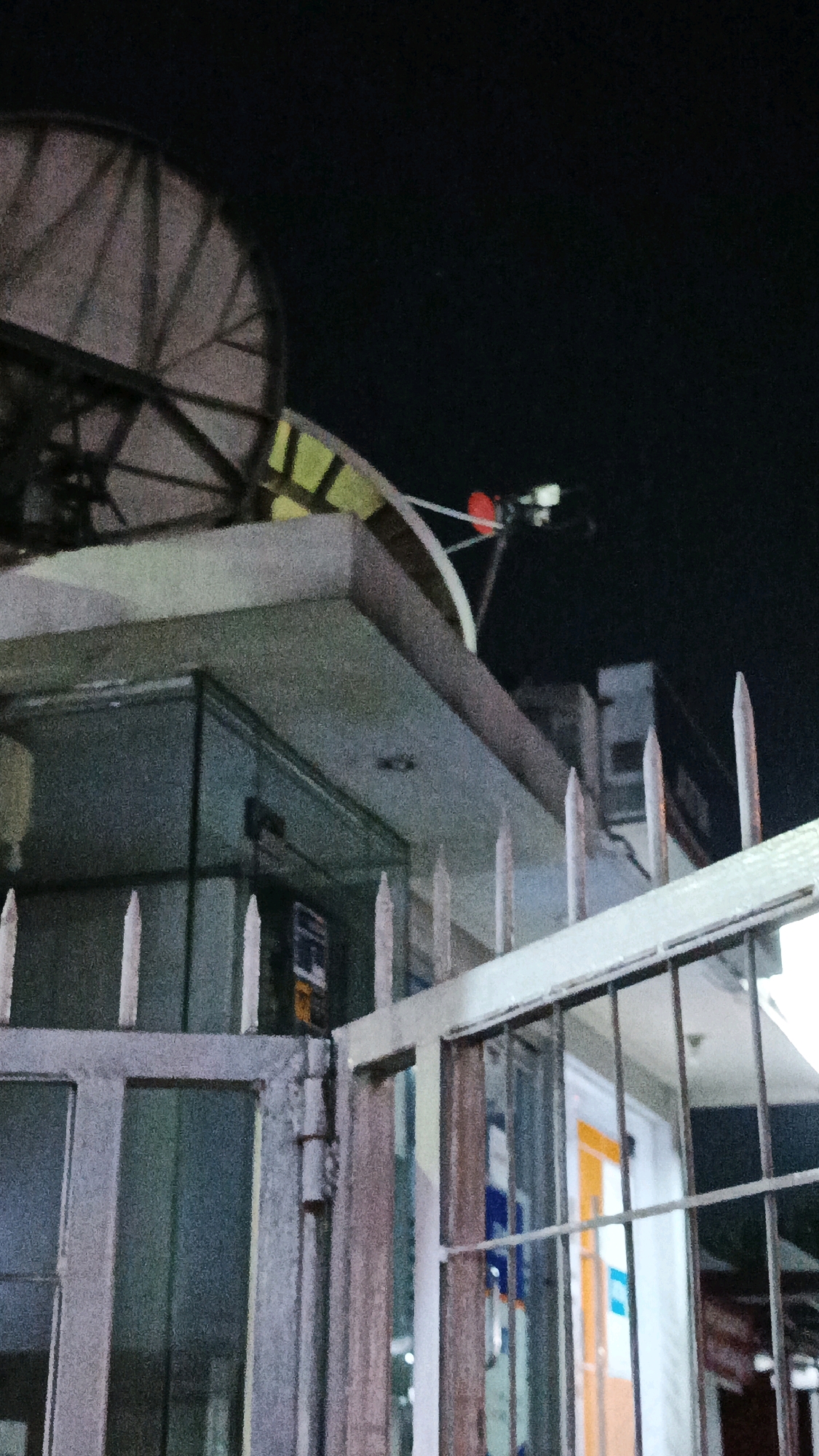 The image shows a night view of a satellite dish mounted on the roof of a building in Surakarta, Indonesia. A spiked fence is in the foreground, with a gate to the left. The building appears to have glass windows, and there are lights on inside. The sky is dark and clear.