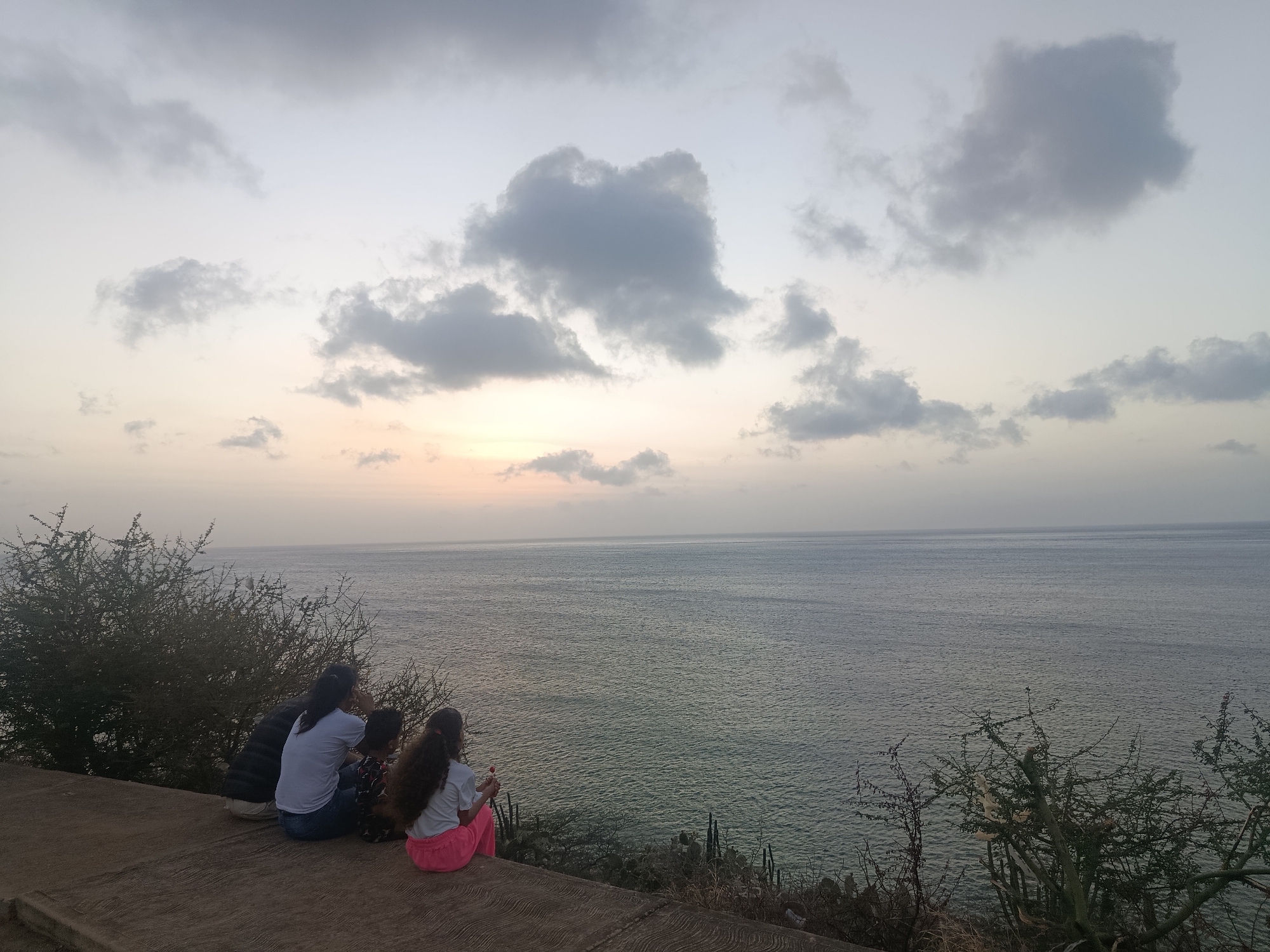The image depicts a scenic view overlooking the ocean in Juan Griego, Venezuela. In the foreground, four people are sitting on a concrete surface, possibly a viewing platform or ledge. They are positioned with their backs to the viewer, facing the water. To the left of the group is a dense bush or shrub, and similar vegetation is visible on the right side of the frame. The sea extends towards the horizon, with a calm surface reflecting the light. The sky is overcast with scattered clouds, and the sun is setting, casting a soft, diffused light across the scene.