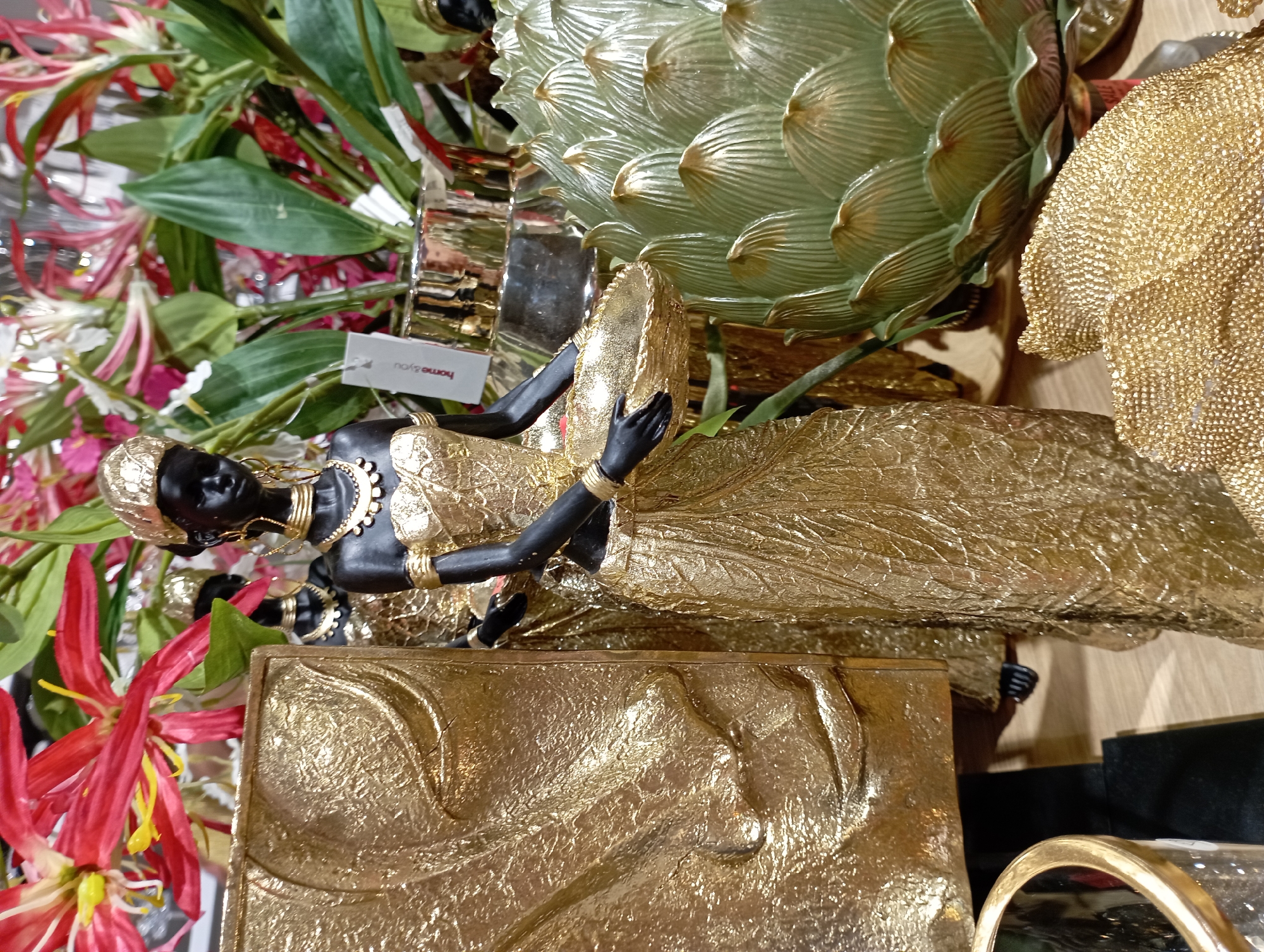 The image depicts a display of decorative items, likely in a store or showroom. In the foreground, a black figurine of a woman adorned in a gold dress and headdress holds a gold bowl. The figurine is positioned to the right of a textured gold rectangular plaque. Behind the main figurine, another similar figurine is partially visible. To the right of the figurines, a large, green, artichoke-shaped decorative object with gold accents is prominently displayed, and to the front right, a golden, textured decorative piece resembling a fan or shell is visible. In the background, artificial flowers with red and white petals, along with green foliage, create a vibrant backdrop. A metallic gold vase is also visible, along with a small white tag with the text "home & you." The overall aesthetic suggests a focus on opulent, metallic decorative pieces with an African-inspired theme. The image does not contain any discernible information that would allow for the identification of its specific location within the city of Katowice, Poland.