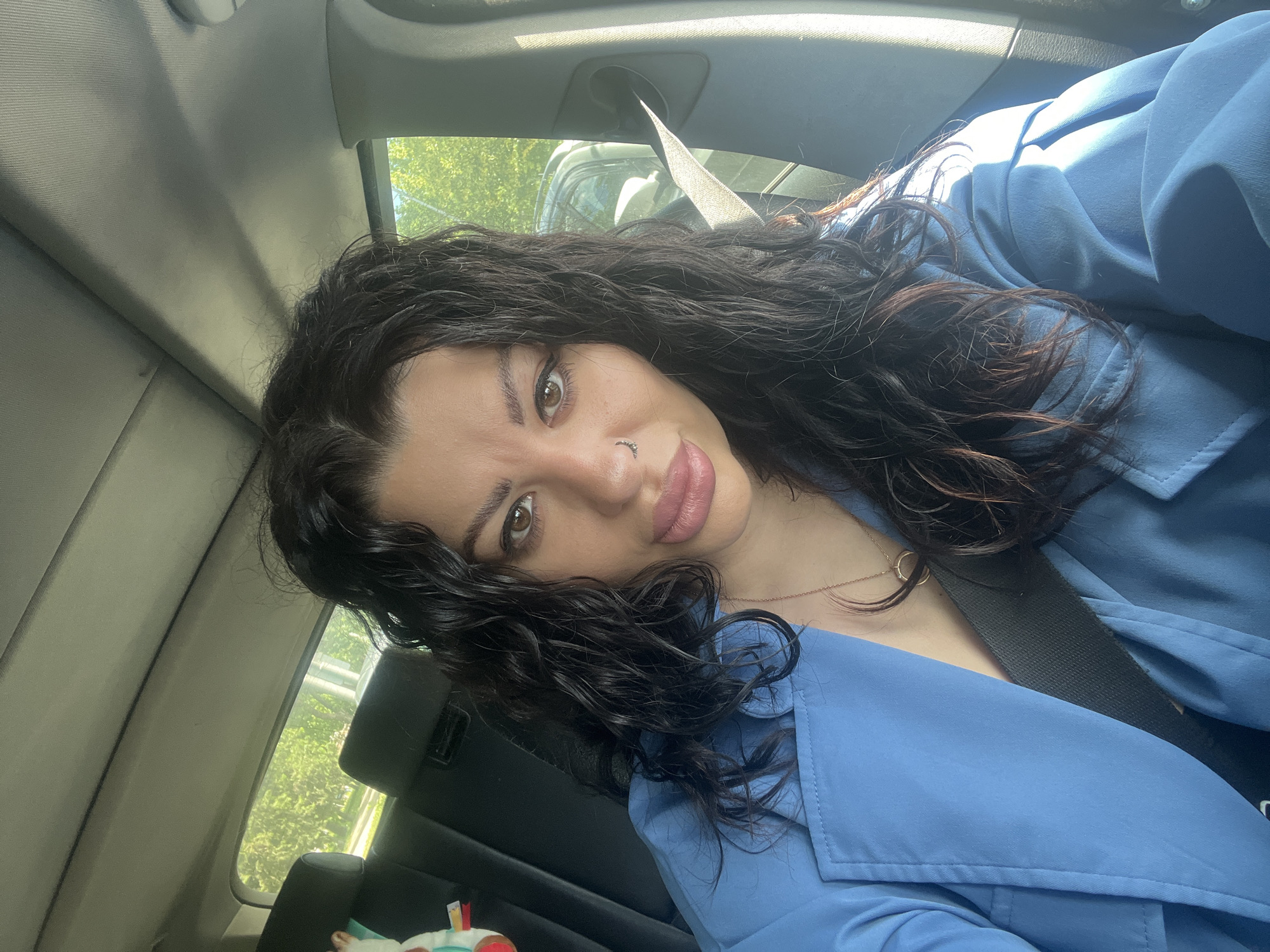 The image shows a woman in a car. She is wearing a blue shirt or jacket and a seatbelt. She has dark, curly hair and is looking at the camera. The background shows a car interior and a glimpse of greenery outside the window. It is not possible to determine the specific location in Plovdiv, Bulgaria from the image.