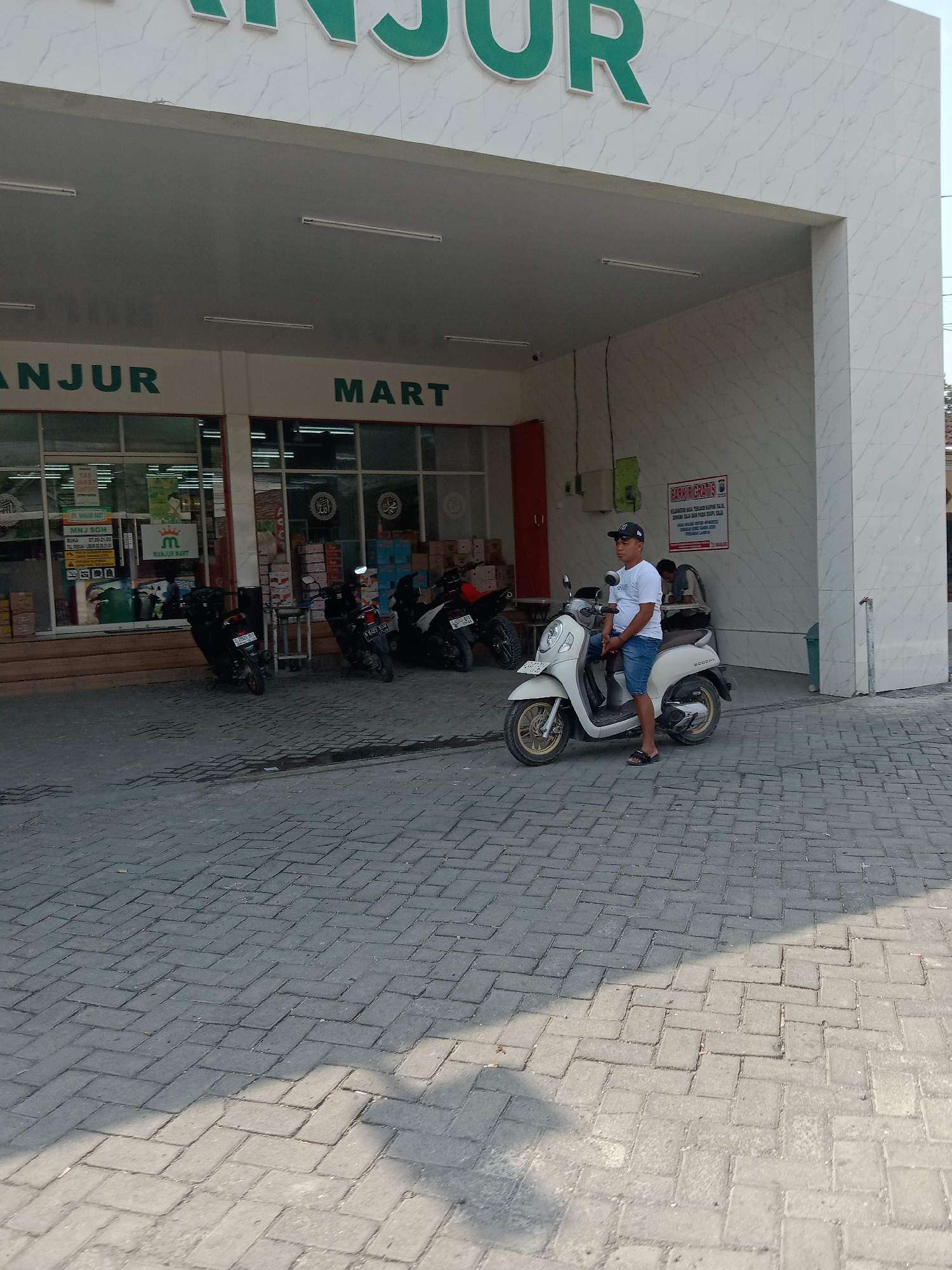 An outdoor, eye-level, full shot captures the entrance of a "MANJUR MART" store, with a man sitting on a white scooter in the foreground. The store entrance is wide and open, with glass doors revealing shelves of goods inside. The building's facade features white marble-patterned tiles and large green lettering that spells out "MANJUR MART." Several motorcycles are parked in front of the store. The man on the scooter wears a white t-shirt, blue shorts, a black baseball cap, and dark sandals. He is looking to his left. The ground is paved with interlocking grey bricks, with a shadow cast by an unseen object to the right. The scene appears to be in Balongrejo, Indonesia, based on the signage.