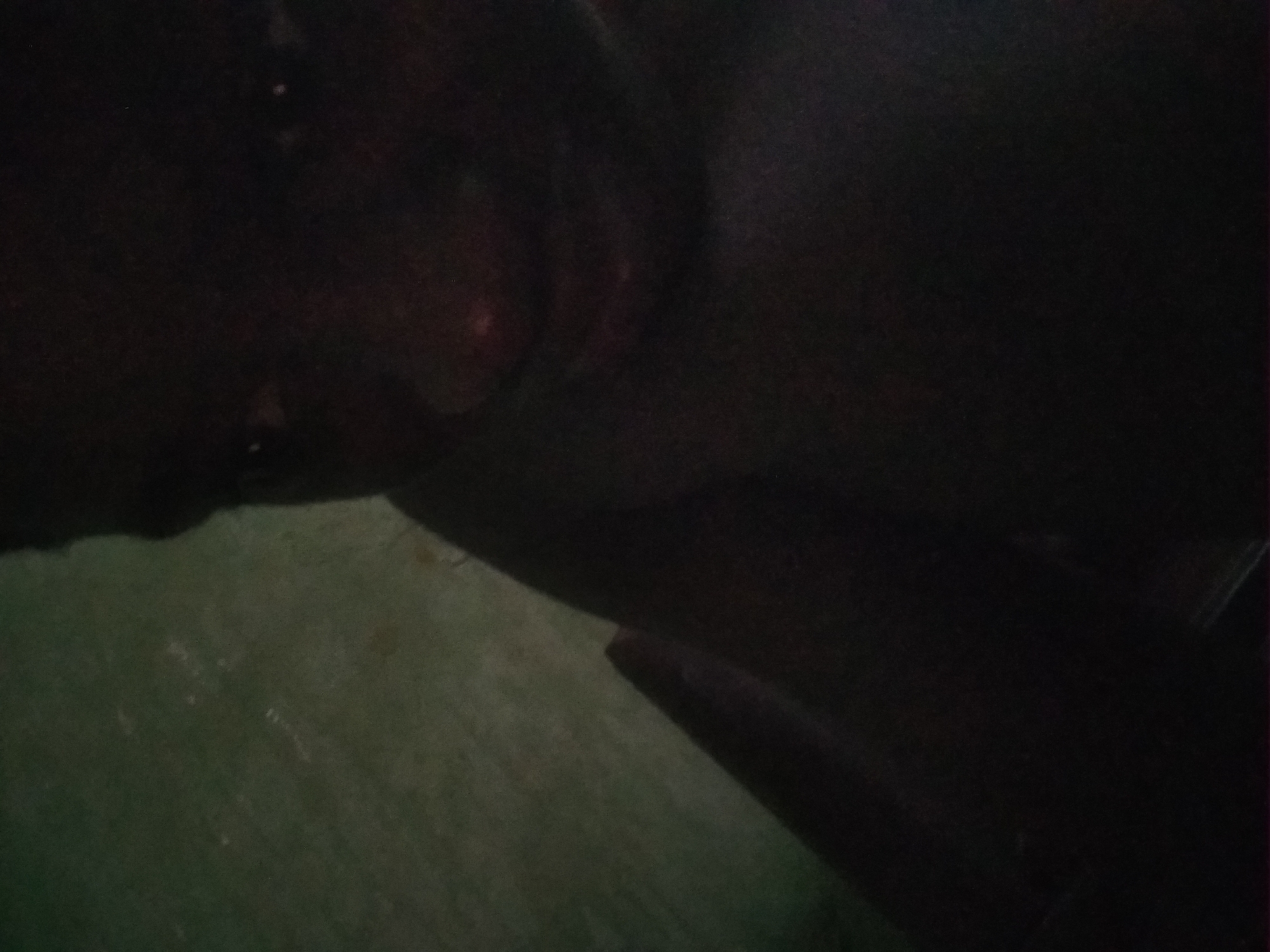 The image is underexposed, showing a person's face and upper body in the dark. The background is a mottled green surface. The location and specific details about the person, objects, and actions are indeterminable from the image provided. The location in Shangisha, Nigeria, cannot be verified.