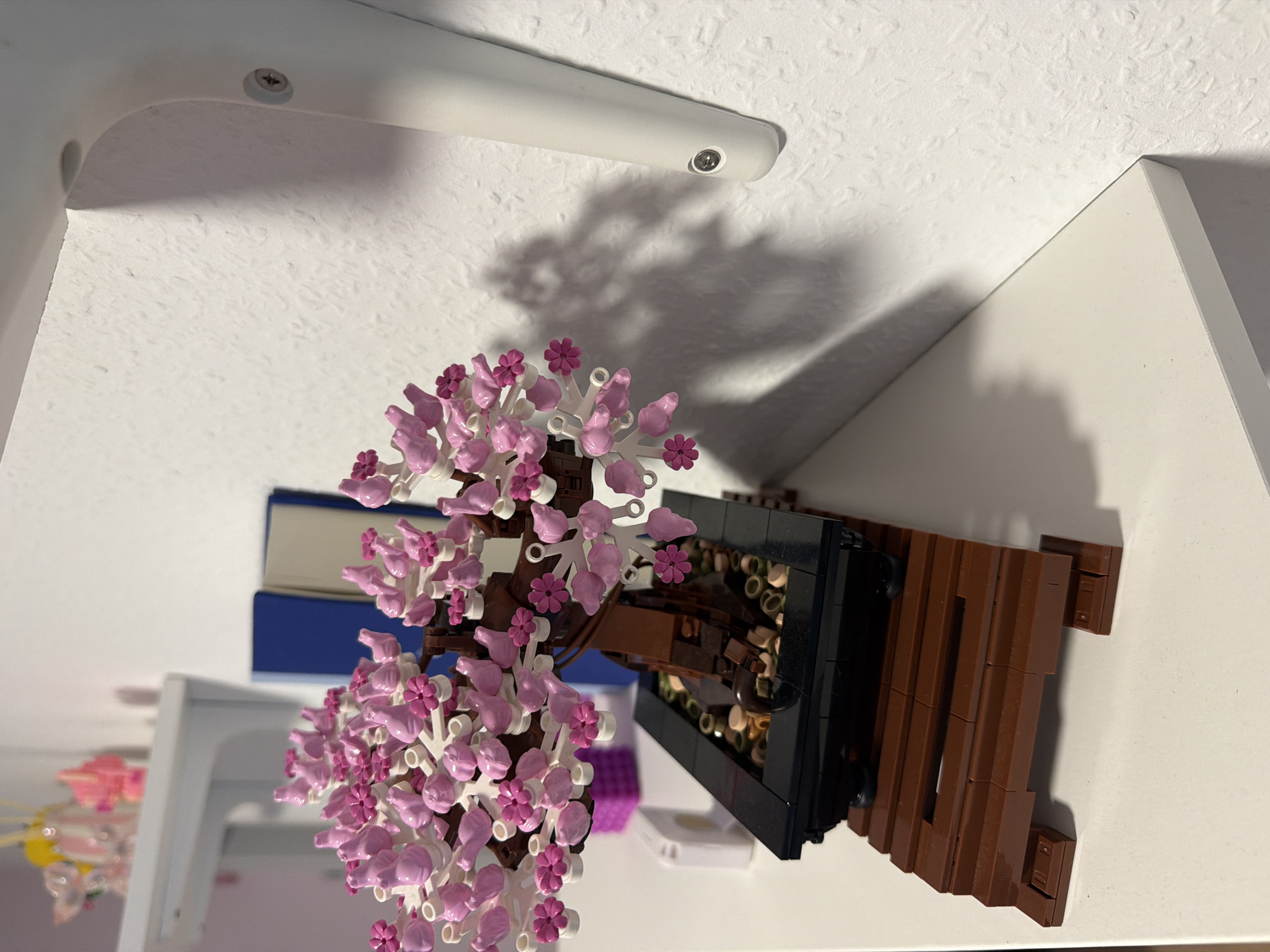 A LEGO cherry blossom bonsai tree sits on a white shelf against a textured white wall. The bonsai has a brown trunk and branches adorned with pink and white blossoms. It is planted in a black pot, which rests on a brown wooden platform made of LEGO bricks. Behind the bonsai, on the same shelf, are two blue and tan books, and further in the background, a small pink decorative item can be seen on a higher shelf. A shadow of the bonsai tree is cast on the wall to the right. The scene appears to be indoors.