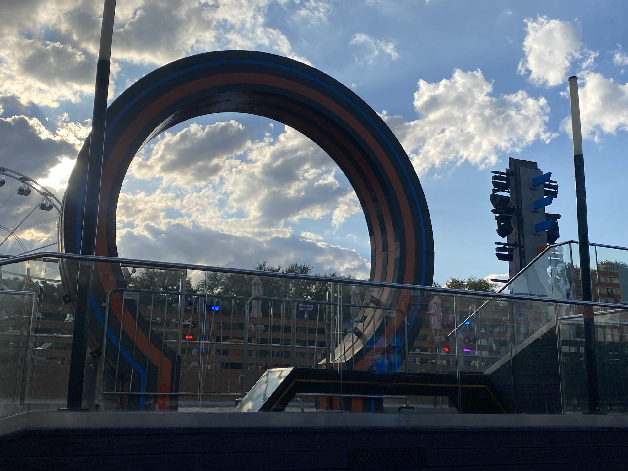 The image depicts a large, circular, modern art sculpture with orange and blue stripes against a backdrop of a partly cloudy sky with sunlight breaking through. To the left, a Ferris wheel is visible. In the foreground, there is a railing and a staircase. To the right, a tall structure resembling a traffic light with multiple blue directional arrows is present. The scene appears to be an outdoor public space or amusement park. The sculpture and its surroundings suggest a contemporary urban environment. The text "ПРОХОД ЗАПРЕЩЕН" (Passage forbidden) is visible on a sign behind the railing. This sign and the context of the Ferris wheel and sculpture suggest the location is likely an entertainment or amusement area. Based on the visual cues and information readily available online about notable public art and attractions in Moscow, Russia, this appears to be the "Cosmos" sculpture located at the VDNKh (Exhibition of Achievements of National Economy) park in Moscow, Russia.
