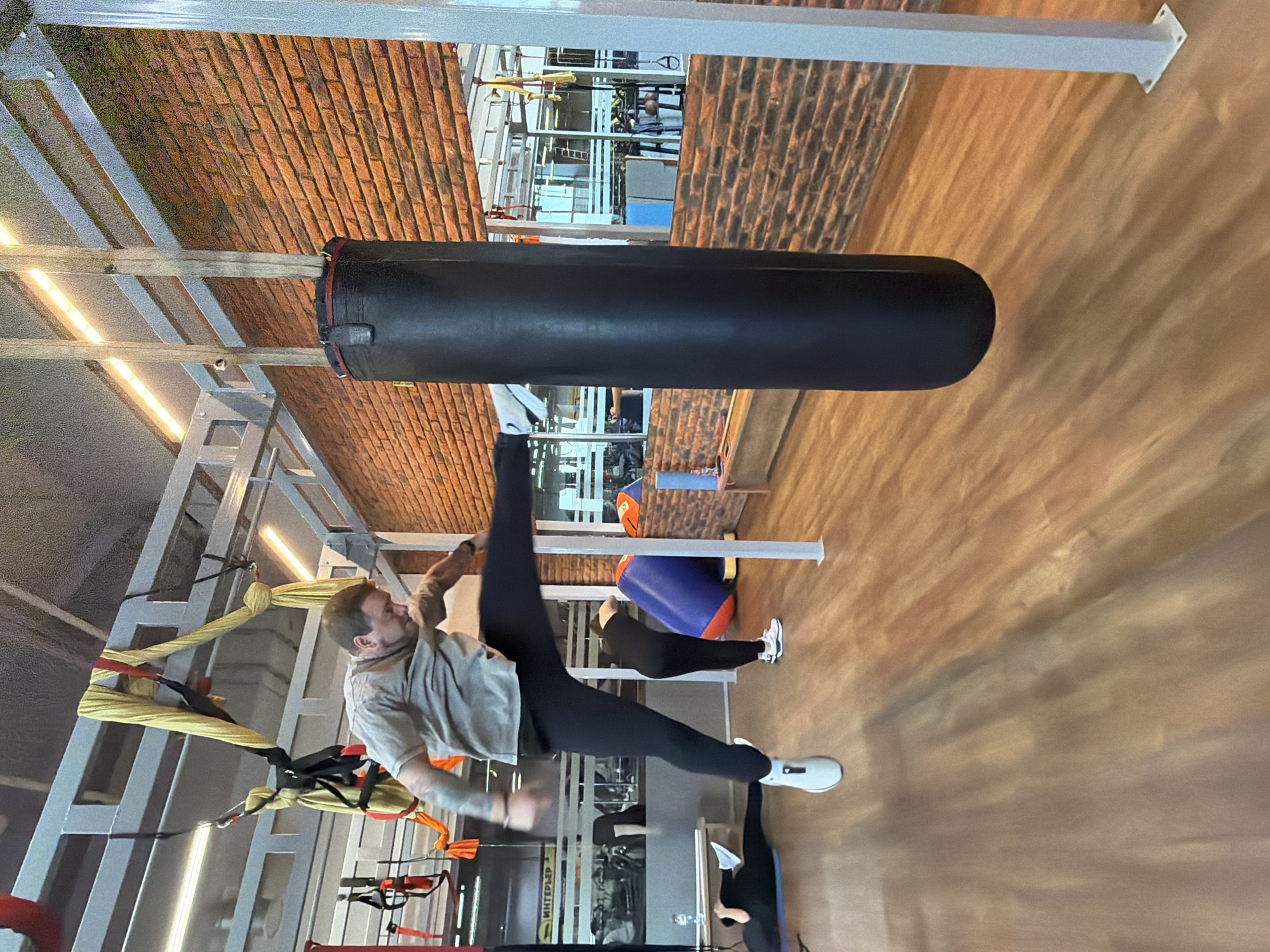 The image captures an individual in a gym setting, likely in the city of Chaikovsky, Russia, given the text on equipment. The individual, a man, is executing a high kick towards a suspended, cylindrical punching bag. He is dressed in a light-colored t-shirt and dark leggings, wearing white athletic shoes.
The gym's infrastructure is visible, featuring exposed brickwork and a structural framework with TRX suspension training equipment. In the background, another person, dressed in black leggings and a light top, is visible near exercise mats and a cylindrical blue cushion. Gym equipment can also be seen in the far background. The floor appears to be wood.