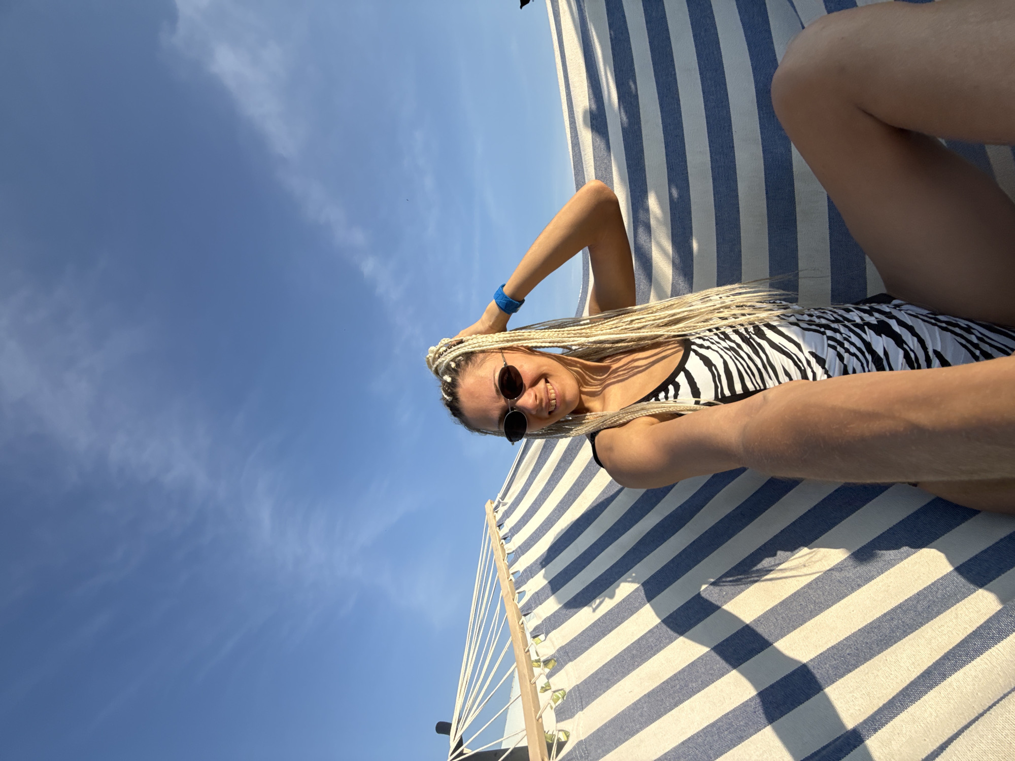 A woman with blonde braids and sunglasses is lying in a hammock. She is wearing a black and white zebra-print swimsuit. The hammock has blue and white stripes. The sky is blue with some clouds. The scene appears to be outdoors, possibly at a beach or park. The location is identified as Rusanovo, Russia.
