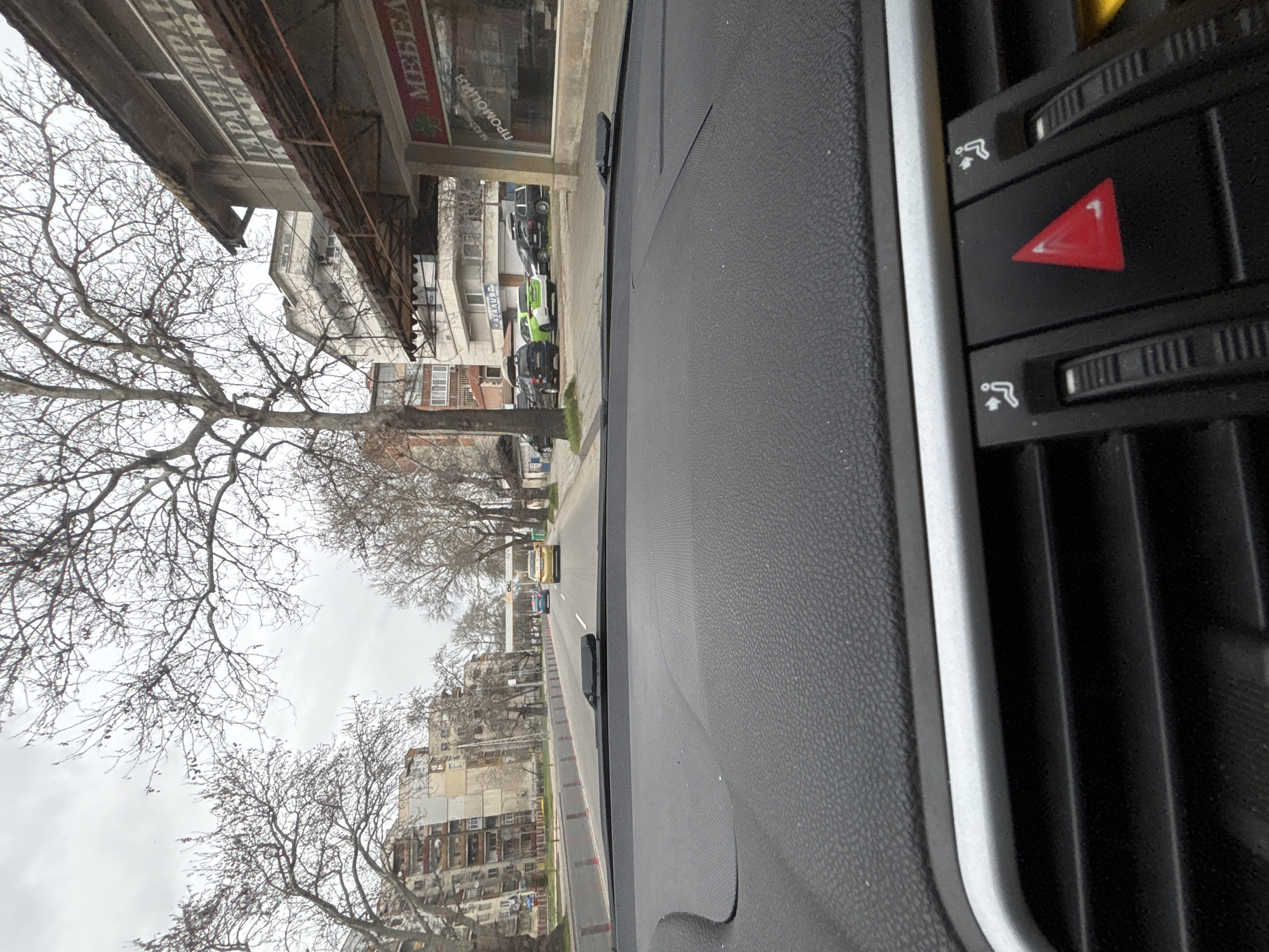 View from inside a car, dashboard and air vents in the foreground. A street scene is visible through the windshield. Buildings with shops are on the right and left sides of the street. Bare trees line the street. Several vehicles are parked along the right side of the road. Cars are on the road ahead. The view appears to be from a street in Plovdiv, Bulgaria.
