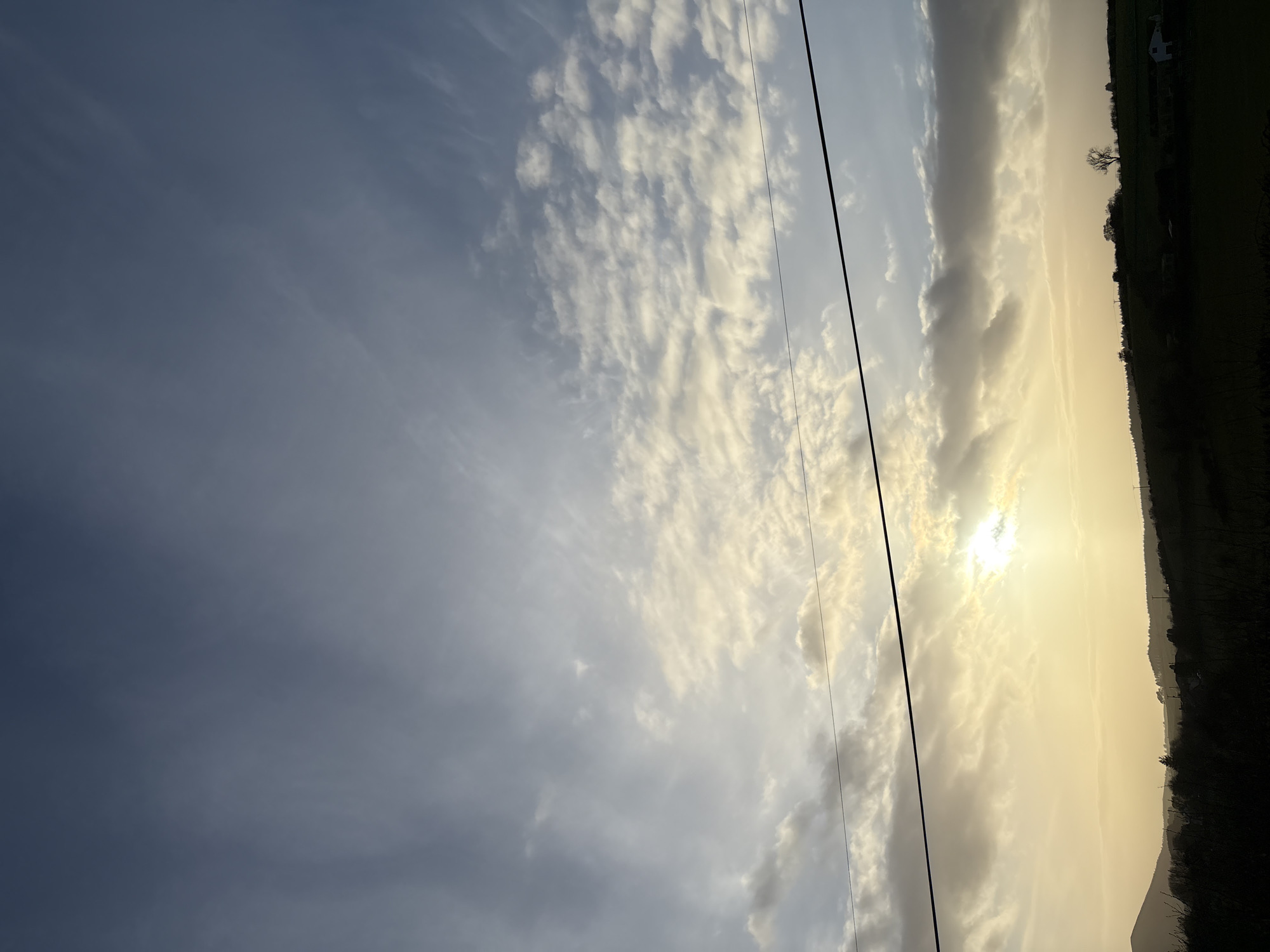 The image depicts a rural landscape at either sunrise or sunset, located near the city of Stiperstones, United Kingdom. The sky occupies approximately the upper two-thirds of the frame, transitioning from a dark blue-grey at the top to a bright yellow-orange and white glow towards the lower-middle where the sun is partially obscured by clouds. Clouds, ranging from scattered altocumulus to more diffuse stratus formations, are present across the sky, particularly concentrated around the sun. A prominent dark utility wire or cable traverses horizontally across the mid-section of the image, positioned above the silhouetted horizon line. The lower third of the image features a dark, silhouetted hilly landscape. Multiple undulating hills and ridges are visible, sparsely populated with trees, some appearing bare. Faint silhouettes of utility poles and at least one structure, possibly a house with a lighter-colored wall or roof, are discernible on the distant hills. The foreground consists of dark, indistinct vegetation, likely fields or shrubbery. No people, animals, or explicit actions are visible within the frame.