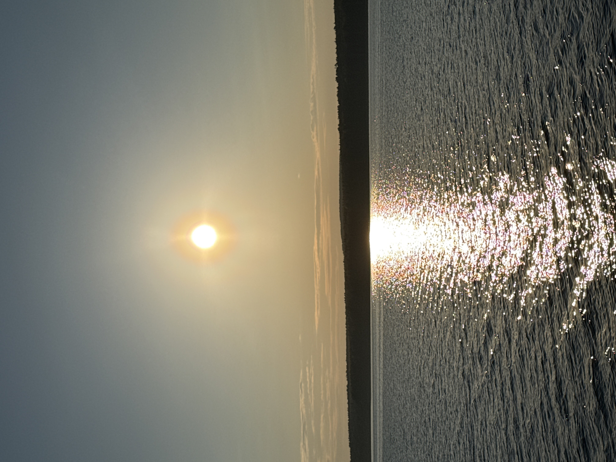 The image depicts a body of water, likely a lake or river, with the sun low in the sky on the left. A strip of land, possibly a forest or shoreline, runs vertically between the water and the sky. The sun's reflection creates a glittering effect on the water's surface. The location is specified as Chaikovsky, Russia, but the image lacks specific details to confirm this, such as visible landmarks or signage. There are no people visible in the scene. The scene captures the interaction of light and water at sunset or sunrise in a natural setting.