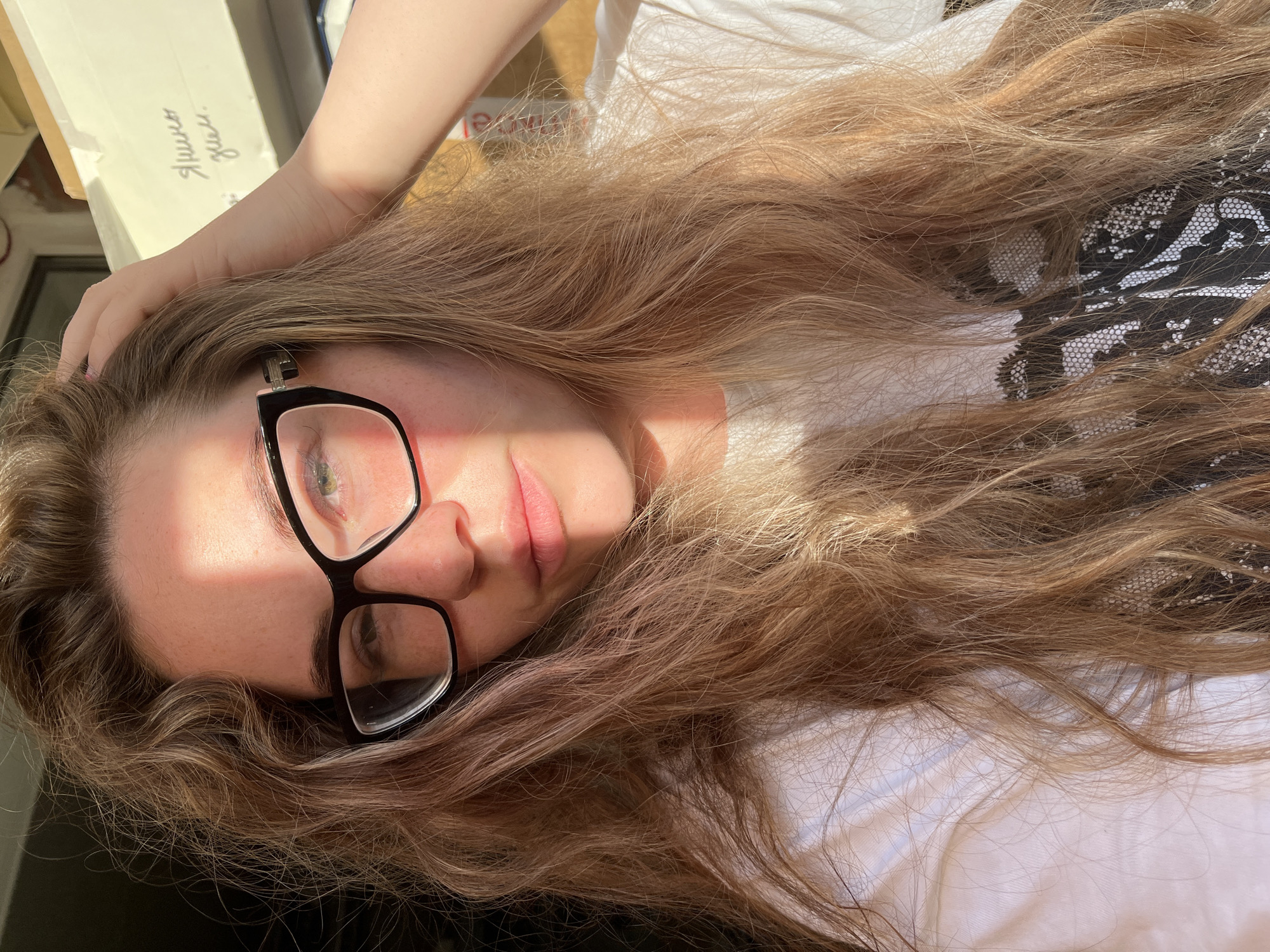A woman with long, light brown hair and glasses is lying down, possibly on a light-colored surface. She has one hand near her head. The image is brightly lit, with strong sunlight creating shadows on her face and the surrounding area. The background is indistinct, with a glimpse of what appears to be a box with Russian writing on it. There is not enough information to confirm the location in the city of Chaikovsky, Russia.