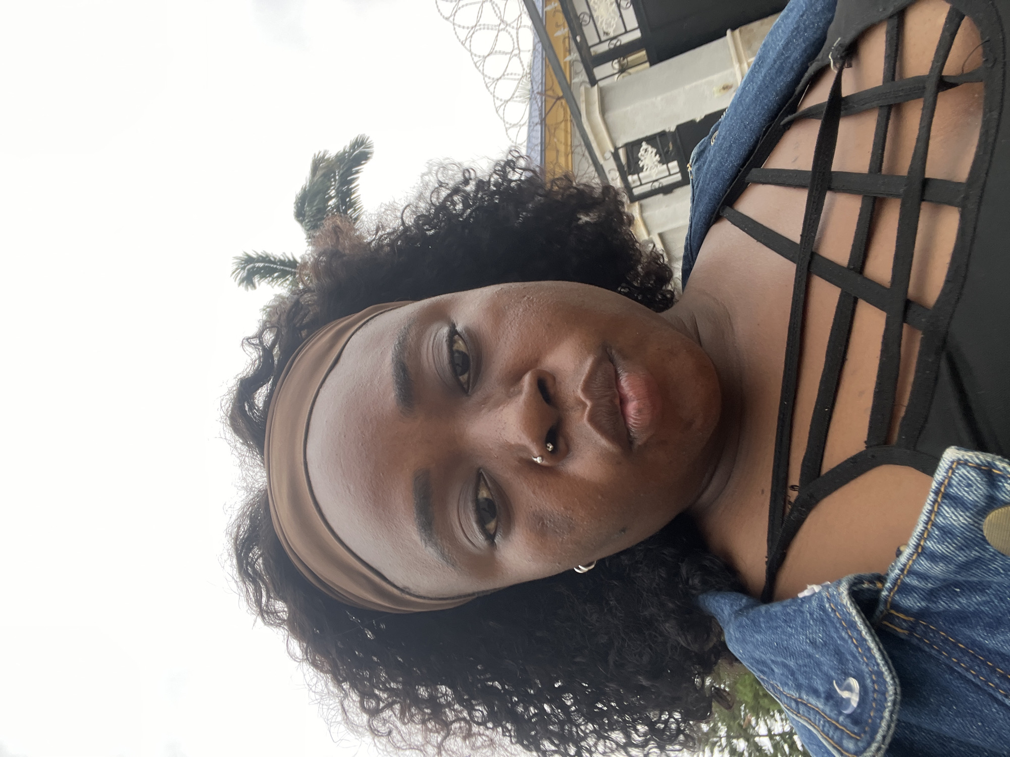 A young Black woman with curly dark hair and a brown headband is looking at the camera with a neutral expression. She is wearing a denim jacket over a black strappy top. She has a nose ring and earrings. The background shows a cloudy sky, a palm tree, and a fence with barbed wire. The location is identified as Abuja, Nigeria.