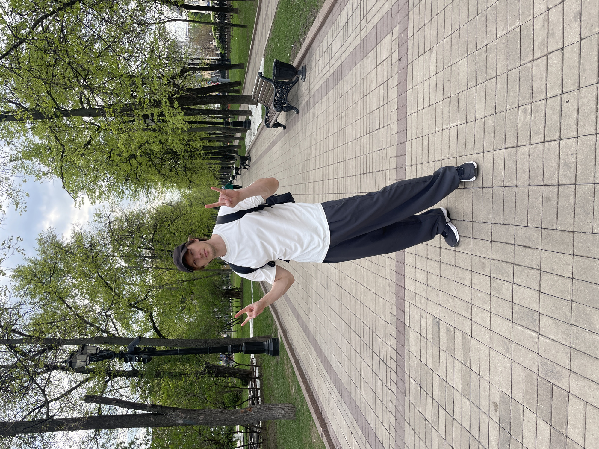 A young man stands on a paved walkway in a park. He wears a white t-shirt, dark pants, a dark cap, and dark sneakers with white soles. He is holding up two fingers on each hand, making a "peace" or "victory" sign. A black bag is slung over his shoulder. The walkway is made of light-colored rectangular stones arranged in a running bond pattern with strips of darker stones. To the left of the walkway, there is a grassy area with trees, benches, and a lamppost. In the distance, more trees and benches can be seen. This is likely a park in Moscow, Russia.