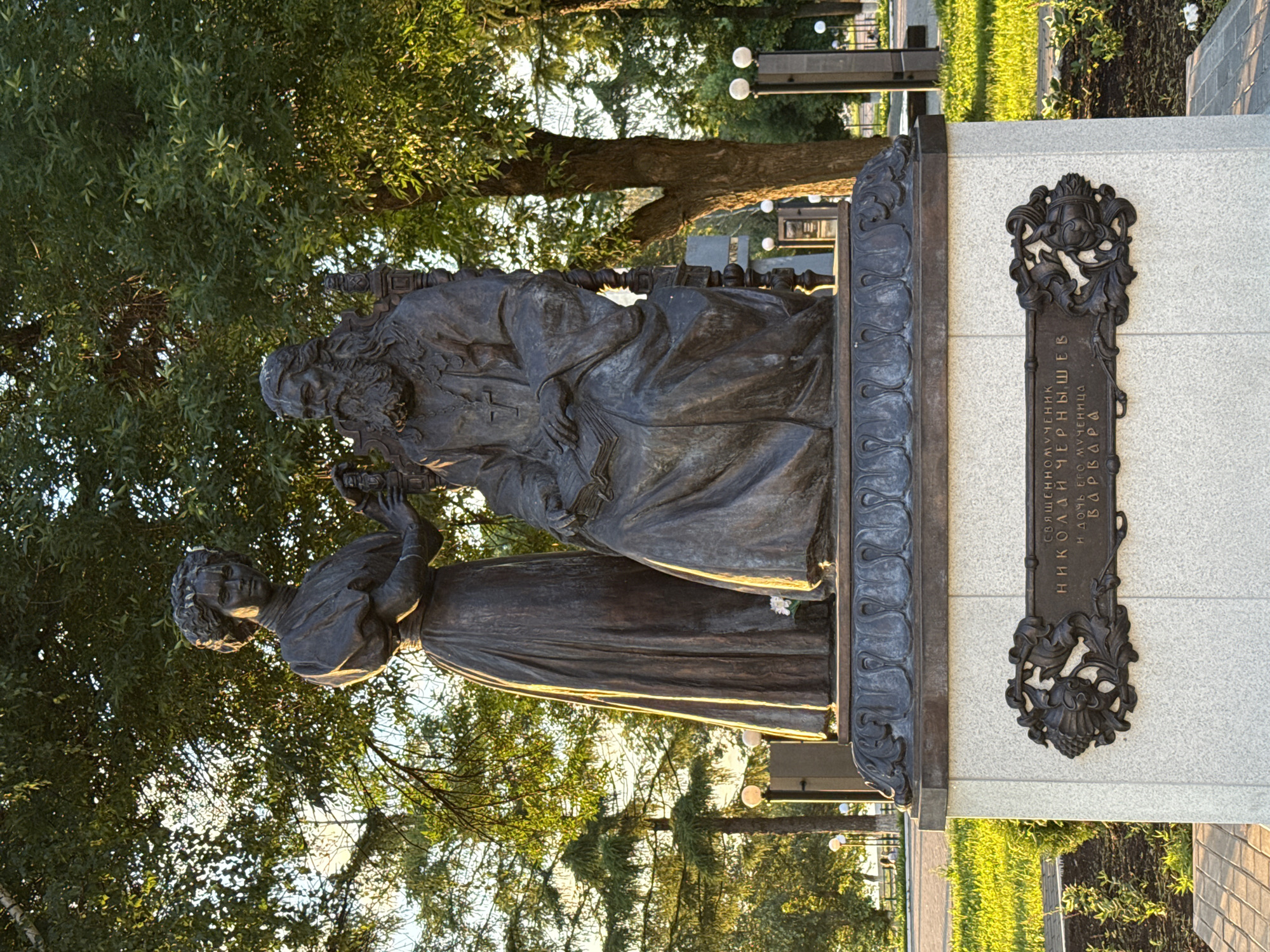 The image shows a bronze monument in the city of Votkinsk, Russia, dedicated to Hieromartyr Nicholas Cherntyshev and his martyr daughter Barbara. The monument features two figures: a seated bearded man in clerical robes holding a book, and a standing woman placing a hand on his shoulder. They are positioned on a white granite pedestal with a decorative bronze plaque inscribed with their names in Russian. The monument is surrounded by lush green grass, trees, and a pathway with lampposts in the background. The scene is bathed in the warm light of a sunset, casting long shadows and highlighting the texture of the bronze and stone.