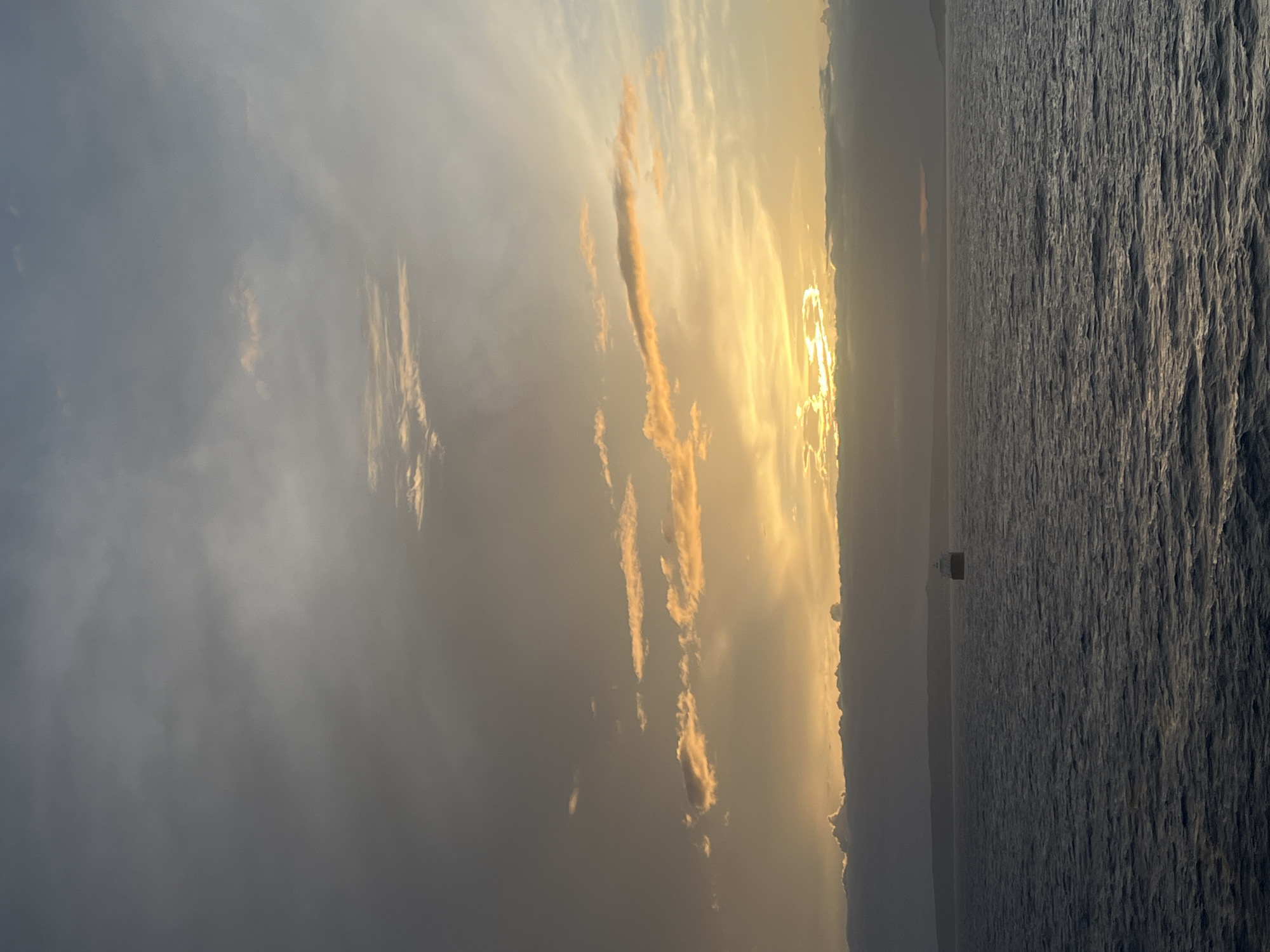 A vertical shot shows a sunset over the ocean. The sky is filled with clouds, some of which are lit up with the golden light of the setting sun. The sun itself is a bright, shimmering disc in the distance, with its light reflecting on the water. The ocean is dark and choppy, with waves visible on the surface. In the distance, a small structure is visible on the horizon line. The location is unknown.