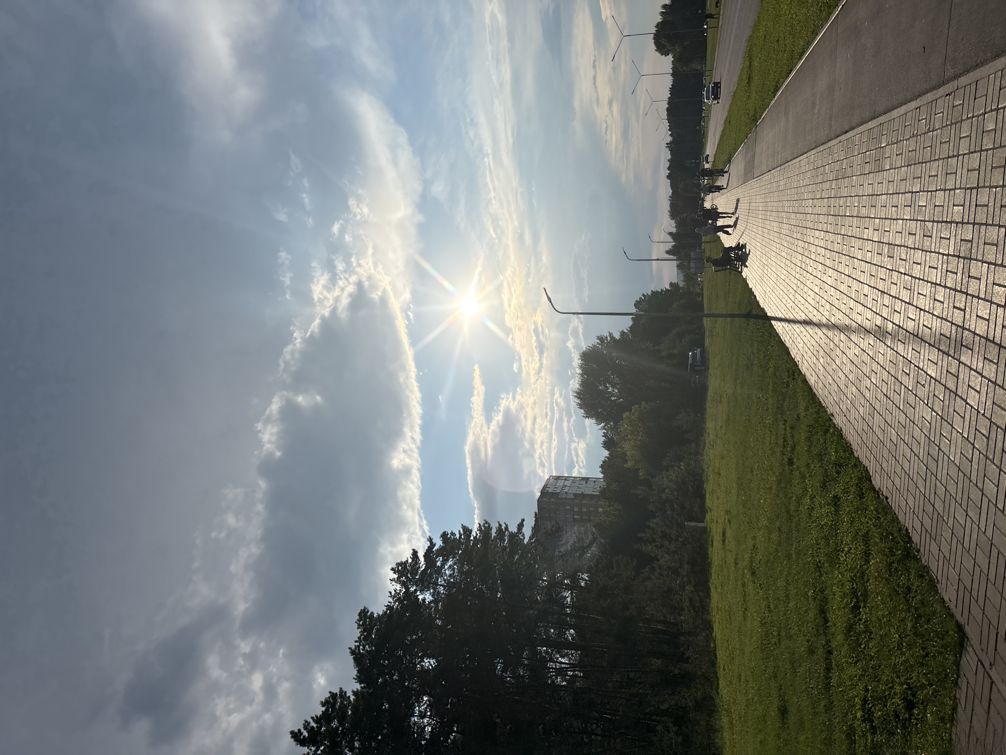 The image captures a sun-drenched, paved pedestrian walkway in Naberezhnye Chelny, Russia, running alongside a grassy verge and a road. The sun is low in the sky, casting long shadows and creating a starburst effect. Several people are visible walking and cycling on the path. A few parked cars and streetlights line the road. In the background, a tall residential building and a dense line of trees are present. The sky is partly cloudy with dramatic cloud formations.