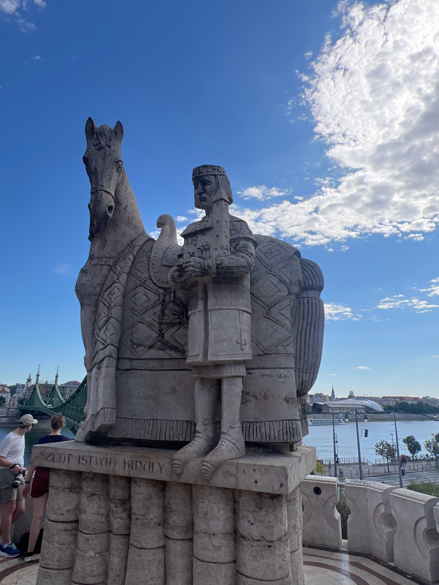The image shows a stone equestrian statue of Saint Stephen, Hungary's first king. The statue is located on Castle Hill in Budapest, Hungary, overlooking the Danube River and the Liberty Bridge. The statue depicts Saint Stephen wearing a crown and armor, holding a sword and a globus cruciger, symbolizing his reign and Christian faith. The horse is depicted with intricate armor, and an eagle is perched on its back. Two people are visible in the foreground, looking at the statue. The sky is blue with scattered clouds, and the overall scene suggests a bright, clear day. The inscription on the base of the statue reads "SZENT ISTVÁN KIRÁLY," which translates to "King Saint Stephen."