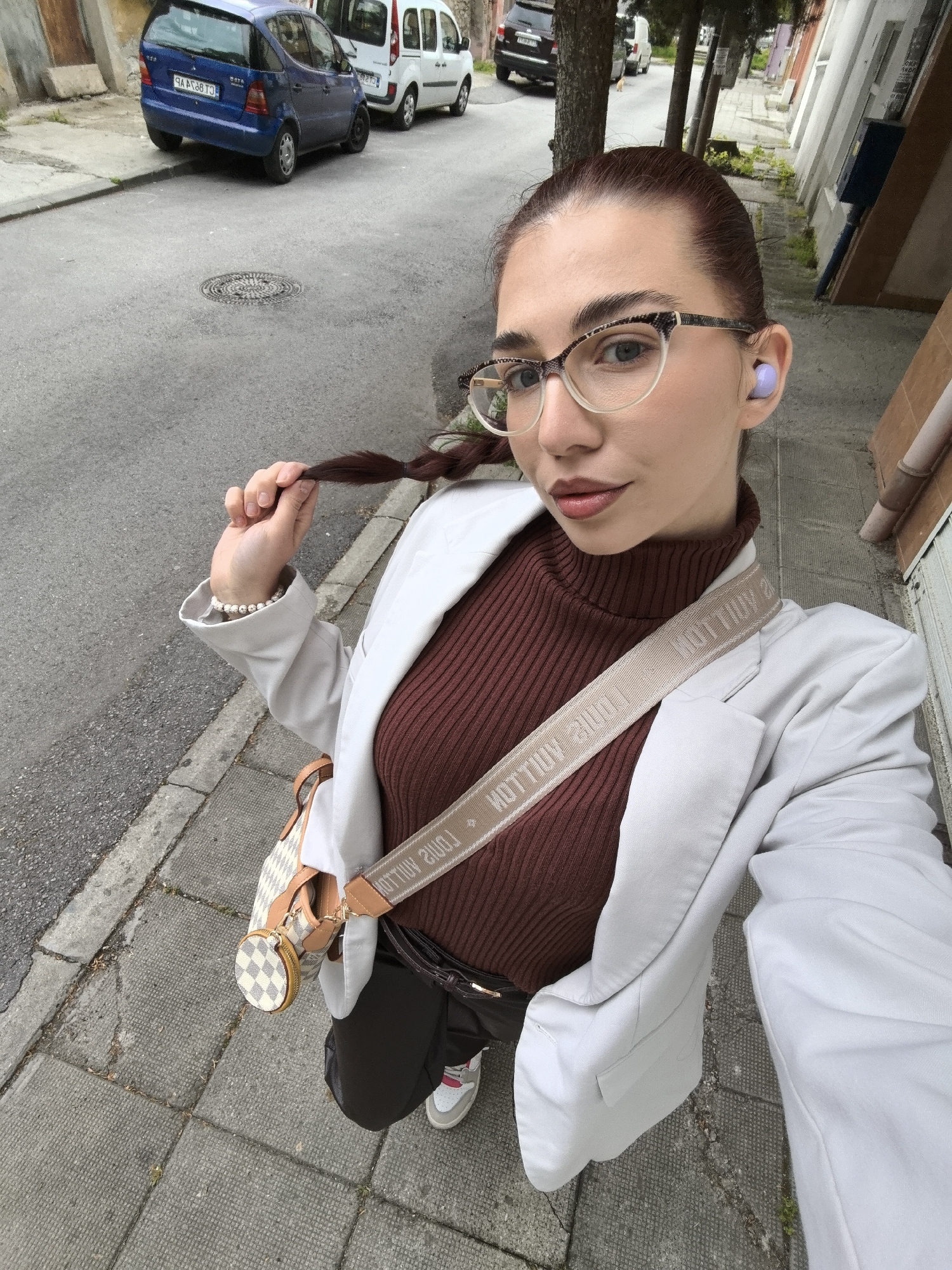 A woman is taking a selfie on a street in Stara Zagora, Bulgaria. She is holding the end of her braided, reddish-brown hair. She is wearing glasses with a snakeskin pattern frame, light purple wireless earbuds, a brown turtleneck shirt, a light gray blazer, and a Louis Vuitton bag. A pearl bracelet is visible on her left wrist. The street has a sidewalk with gray paving stones. Behind her, there is a blue car, a white van, and a dark SUV parked on the street. Buildings with white facades line the other side of the street. There are trees along the sidewalk as well. The sky appears overcast. A manhole cover is visible in the street.