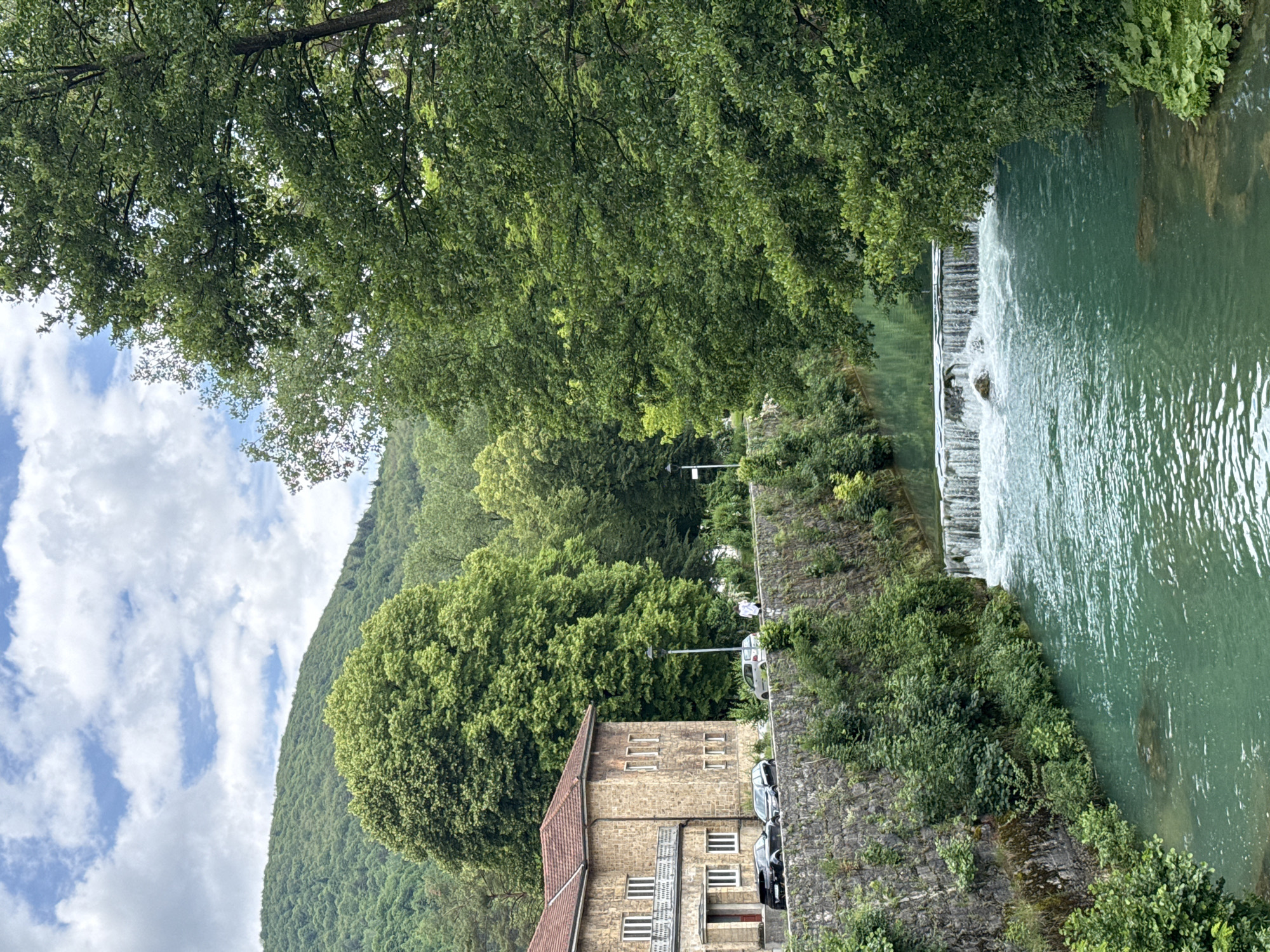This image captures a view along a river in the city of Jajce, Bosnia and Herzegovina, under a sky with prominent white cumulus clouds against patches of blue. The central feature is a wide river, exhibiting a distinct turquoise-green coloration, flowing from the background towards the foreground. A low, linear weir or waterfall spans the river approximately mid-frame, over which white water cascades and froths around visible rocks.
The left bank of the river is defined by a multi-tiered stone retaining wall, constructed from irregular blocks and partially covered in green vegetation and moss. Above this wall, a paved road or pathway runs parallel to the river. Three automobiles are parked on this road: a dark SUV, a black sedan, and a silver sedan. Two dark-colored streetlights are positioned along the road. Adjacent to the road, on the far left, a two-story building with a reddish-brown tiled roof and textured stone walls is visible. It features multiple rectangular windows with white frames and a second-story balcony with an iron railing. A red door is present on the ground level. A single individual, dressed in a light-colored, long garment, walks on the road away from the building.
The right bank of the river is characterized by a dense growth of mature, deciduous trees with lush green canopies that extend into the frame and undergrowth reaching the water's edge. In the background, a large, densely forested mountain rises, covered in continuous green foliage, under the same partly cloudy sky. The scene integrates natural riverine and mountainous topography with urban elements of a road, parked vehicles, a building, and streetlights.
