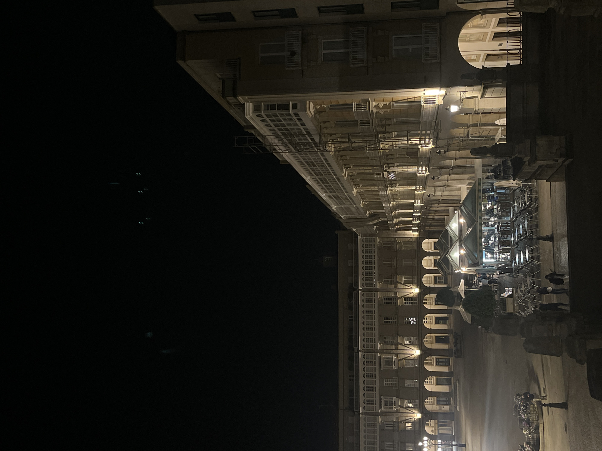 The image depicts the arcaded galleries along the waterfront of A Coruña, Spain, at night. The architecture features multi-story buildings with rows of windows and arched porticos. Lights illuminate the galleries, and there is outdoor seating set up, with some people present. There are reflections on the ground that indicate wet conditions. The night sky is dark.