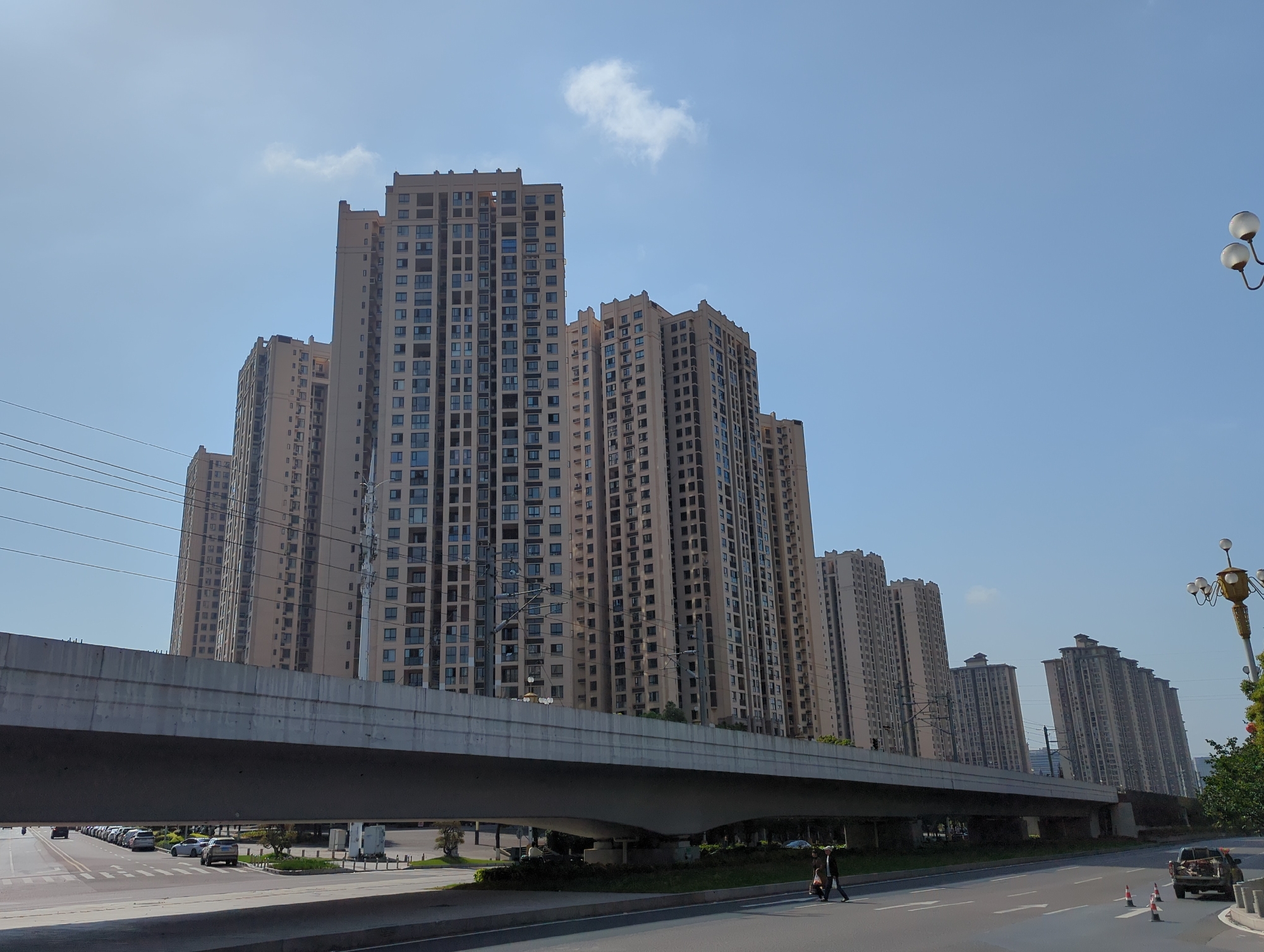 This image depicts a modern, densely populated urban environment in Jiangjin, China, characterized by numerous high-rise residential buildings and extensive infrastructure.
**Subjects:** The dominant subjects are several tall, light-colored apartment buildings with numerous windows, stretching into the clear blue sky. An elevated concrete overpass or bridge cuts across the middle ground, separating the buildings from the street level. Below the overpass, a multi-lane road is visible, with several cars parked on the left and others driving. A person on a scooter or motorcycle is traveling on the right side of the road. Further right, two individuals are walking on what appears to be a crosswalk or sidewalk, and a pickup truck is seen near traffic cones. Streetlights, some with double spherical lamps and others with more ornate, gold-colored designs, are positioned along the road. Numerous power lines are also visible, especially on the left side of the frame, crisscrossing the sky.
**Activity or event taking place:** The scene shows typical urban life with vehicular traffic and pedestrians. Cars are either parked or in motion, and a scooter rider and pedestrians are navigating the street.
**Notable details:** The image captures a bright, sunny daytime scene under a clear blue sky, with only a few small, white clouds visible. The overall weather appears fair. The architecture of the buildings suggests a contemporary urban landscape. The elevated overpass indicates a complex transportation system. No specific emotional cues are discernible, but the scene is active and functional, typical of a bustling city.
**Visible text:** No discernible text is clearly visible on any buildings, signs, or vehicles in the image.