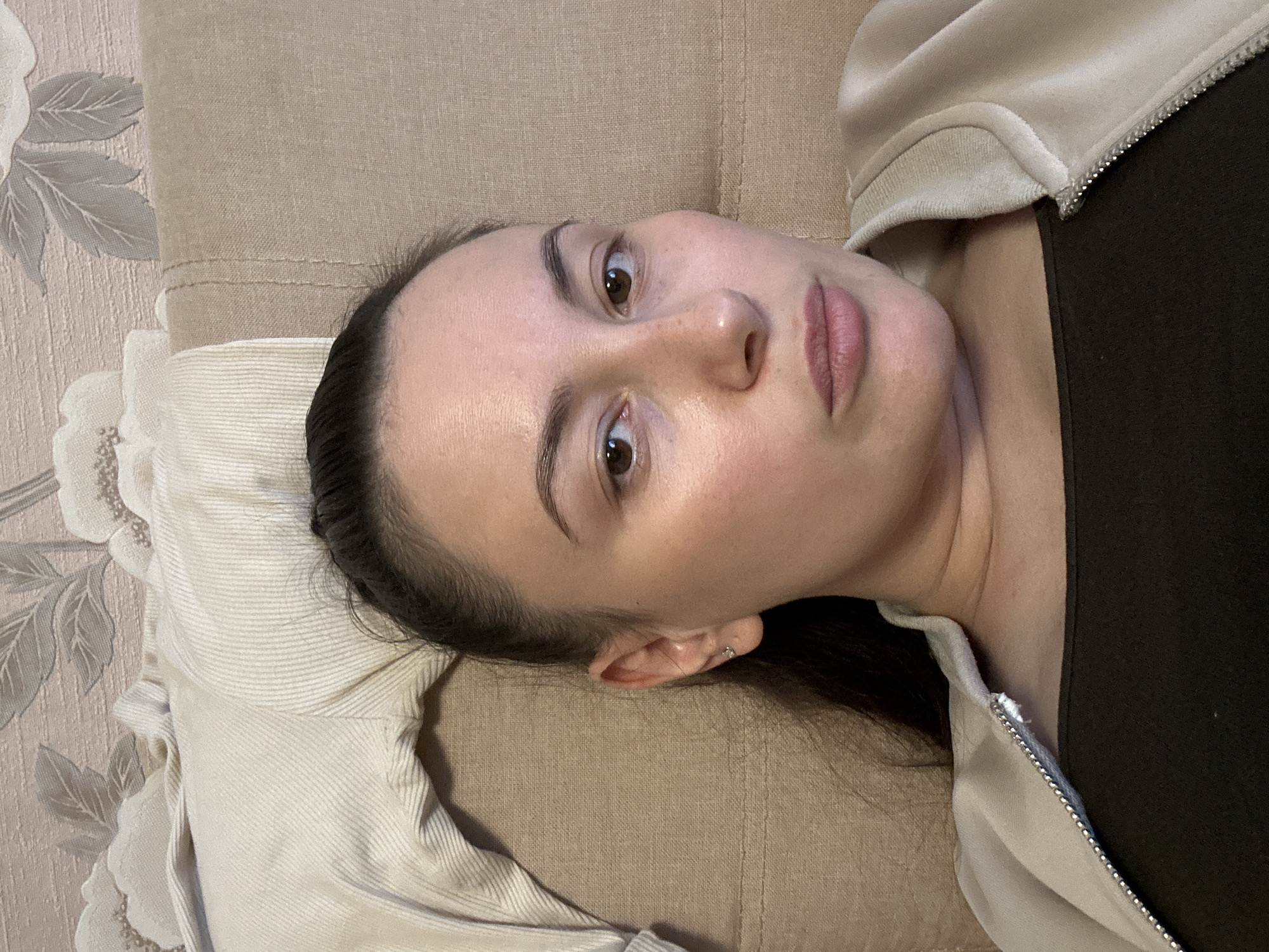 A woman lies on a tan surface, possibly a couch or bed, with her head resting on a patterned pillow. She has dark hair pulled back, brown eyes, and fair skin. She is wearing a dark top and a light-colored zip-up hoodie. The location is not discernible from the image.
