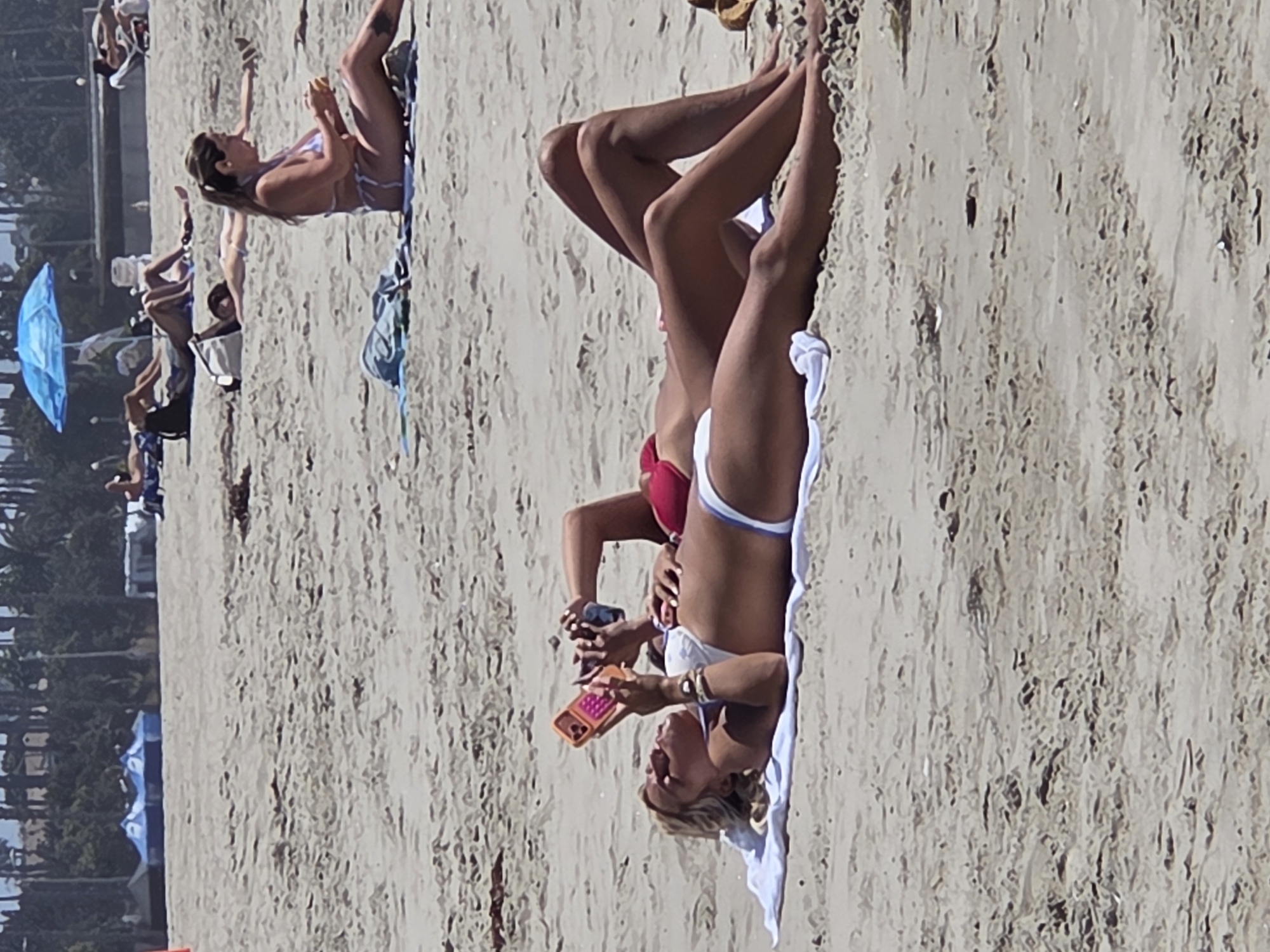The image shows a sunny day at a sandy beach in Santa Monica, California. In the foreground, two women are lying on their backs on a white towel. The woman closer to the camera, on the left, is wearing a white bikini and is holding an orange cell phone. She has blonde hair and is looking at her phone. The woman behind her, to the right, is wearing a red bikini top and her legs are visible. They appear to be relaxed and enjoying the beach. In the background, other people can be seen sunbathing, and there are some blue tents and a blue umbrella on the beach. Palm trees line the back of the beach, and some buildings are visible behind the trees. The sand is light brown and textured with footprints and small ripples. The water is not visible in the frame.