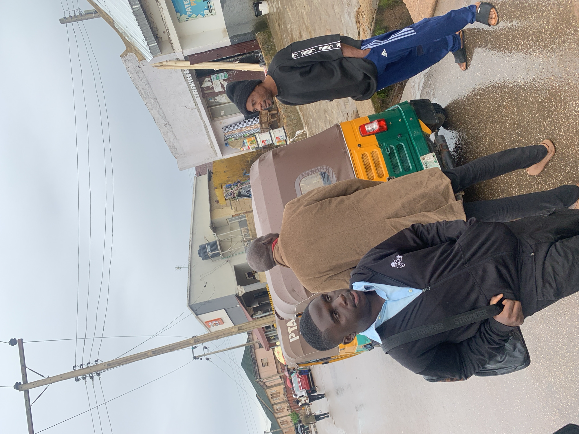 A group of people are standing on a wet, paved street in Jos, Nigeria, next to a yellow and green auto-rickshaw. The sky is overcast. In the foreground, a young Black man wearing a light blue collared shirt and a black zippered hoodie with a backpack slung over his shoulder is looking to his right, with his mouth slightly open. Behind him and to his right, another Black man in a brown coat is facing away from the camera. To the right of the auto-rickshaw, a Black man wearing a black beanie, black sweatshirt, and blue athletic pants with white stripes is looking to his left, with his hands in his pockets. In the background, buildings line the street, with a power pole and wires overhead. The ground is reflective, indicating recent rain.