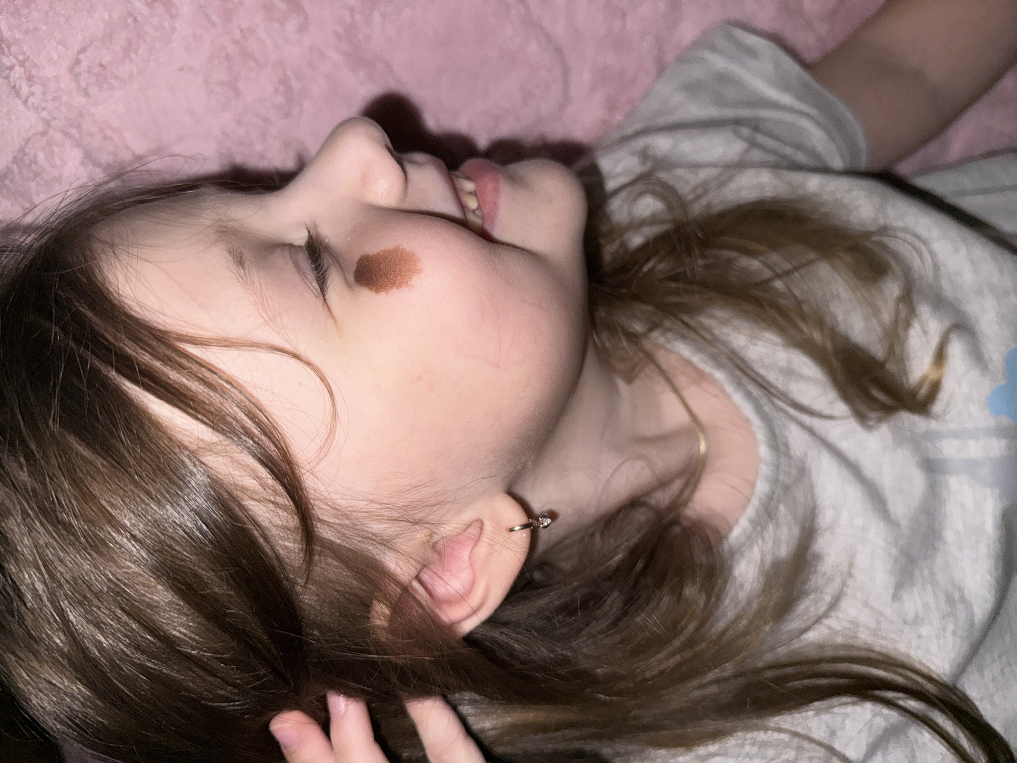 A young girl is lying on her side on a pink surface, possibly a bed. She has light skin, brown hair, and a visible mole on her cheek. She is wearing a grey t-shirt and an earring. The girl is smiling slightly with her eyes closed.
I am unable to identify the location in the city of Кипчак, Russia, as the image does not provide any visual clues related to that location.