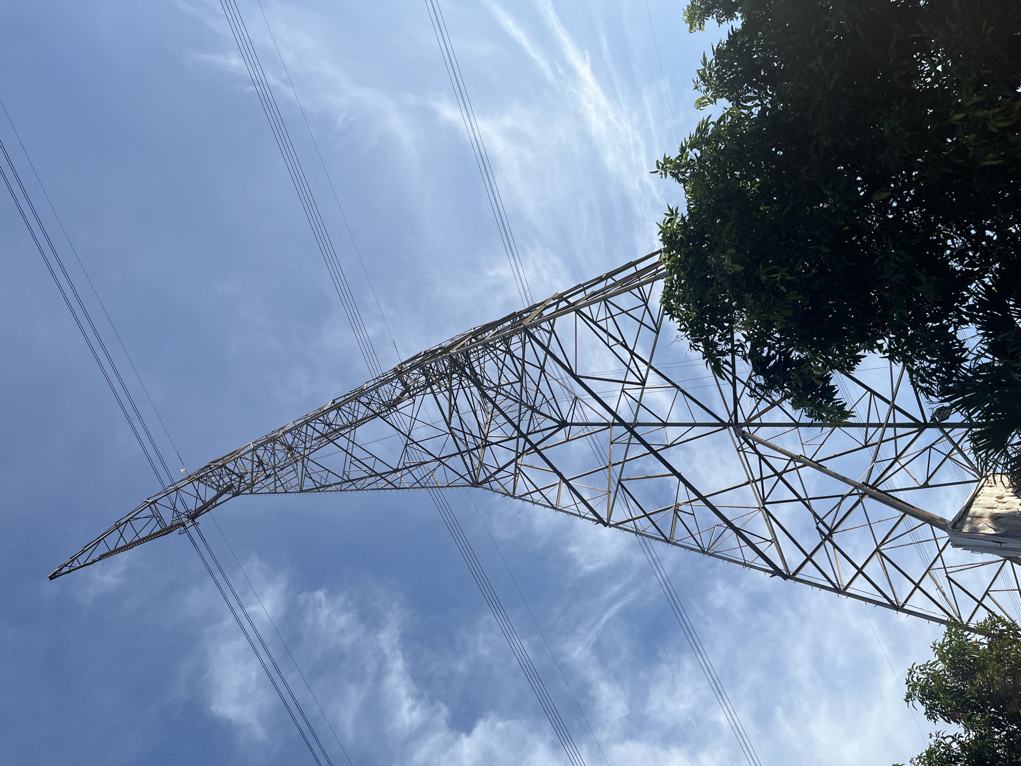 The image shows a low-angle view of a metal electricity pylon against a sky with wispy clouds. The pylon is constructed of a lattice of steel beams, tapering as it extends upwards. Power lines run diagonally across the frame. Tree foliage is visible on the right side of the image. The location is Sukabakti, Indonesia.