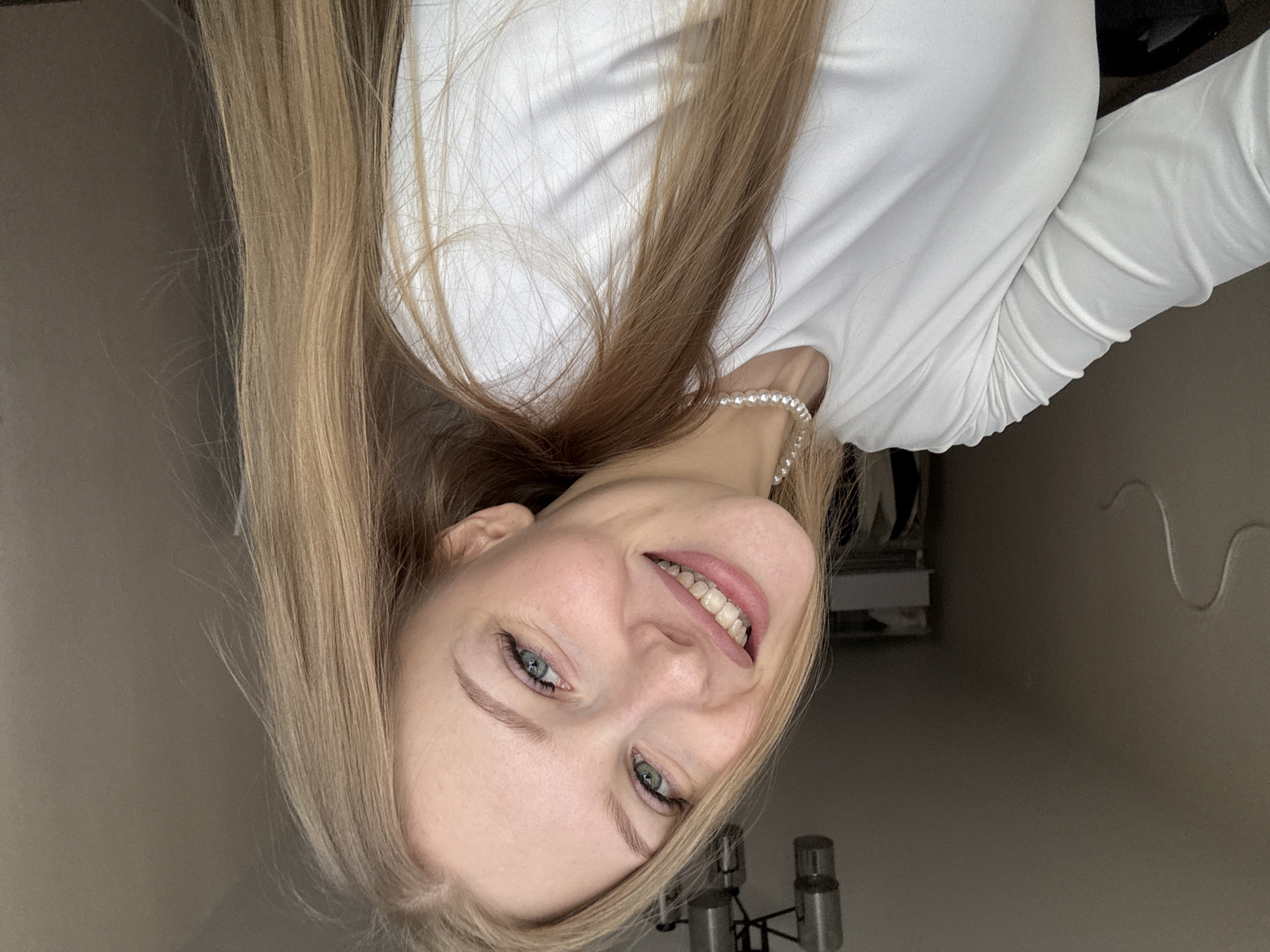 A young woman with long, straight, light blonde or light brown hair is the central focus of this indoor portrait. She appears to be in her late teens or early twenties, with fair skin and expressive blue-grey eyes. Her face is turned towards the viewer, and she sports a wide, open-mouthed smile, revealing her teeth and conveying a cheerful and friendly demeanor.
She is dressed in a simple yet elegant white long-sleeved top, which has a smooth, fitted texture and some subtle ruching or gathering visible on the sleeves. A delicate pearl necklace adorns her neck, adding a classic touch to her attire. The image is captured from a slightly low angle, and her head is tilted, giving the impression that she is looking playfully at the camera.
The background is minimalist, featuring a plain, light-colored wall, which is likely a shade of beige or light grey. In the background, particularly visible in the lower right, is part of a contemporary light fixture, possibly a chandelier, with metallic elements and multiple light sources. The setting suggests a casual home environment. The lighting is even, typical of indoor artificial or diffused natural light, without indicating a specific time of day. No text is visible in the image. The overall impression is one of warmth and approachability.