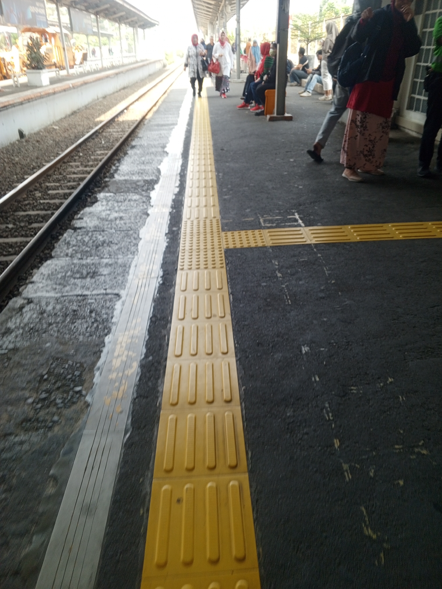 The image depicts a train platform at Depok Station, Depok, Indonesia. Several individuals are present on the platform. In the foreground, a tactile paving strip, commonly known as a braille block, runs along the edge of the platform. The train tracks are visible to the left, with gravel ballast. In the background, people are seen walking and waiting. The platform roof structure and overhead lighting are also visible. The overall lighting suggests it is daytime, possibly morning or late afternoon due to the warm tones.