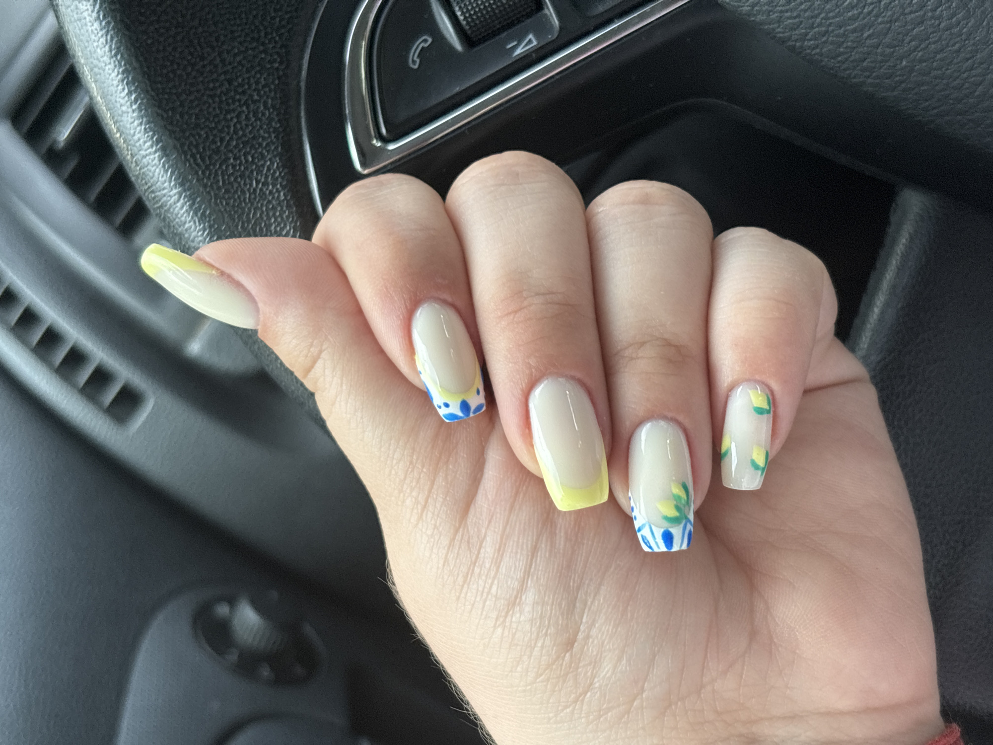 A close-up captures a person's left hand with distinctively decorated fingernails against the backdrop of an automobile's interior. The nails are a pale beige, with a yellow French tip on the thumb and ring finger. The remaining nails are adorned with blue and green floral designs. The hand is positioned in front of a dark, textured dashboard and steering wheel, complete with control buttons. The location of the photograph is not in the city of Plovdiv, Bulgaria. The photograph lacks any details of Plovdiv, Bulgaria.