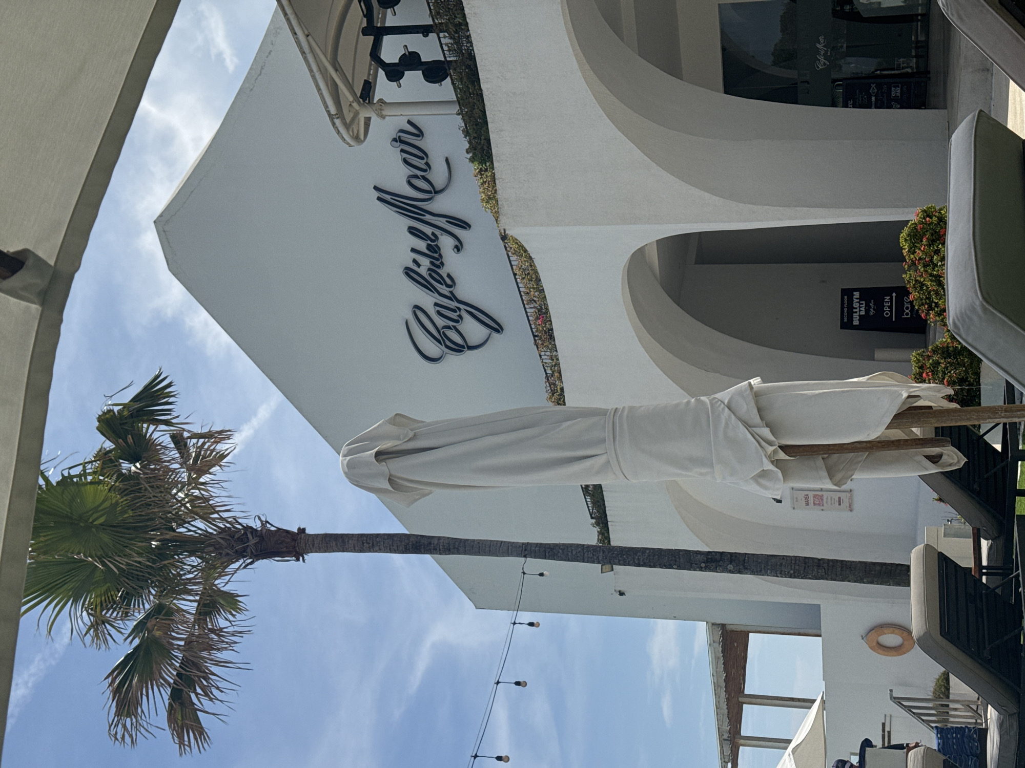 The image shows the Cafe del Mar in North Kuta, Indonesia. The architectural style is modern, with white rounded structures. The cafe's name, "Cafe del Mar," is displayed prominently on the building. A palm tree extends toward the sky on the left side of the frame. A closed white umbrella stands next to outdoor furniture. String lights hang above. A sign reading "SECOND FLOOR BULLGYM BALI CafeGym" is visible, along with an "OPEN" sign. The sky is partially visible with light cloud cover.