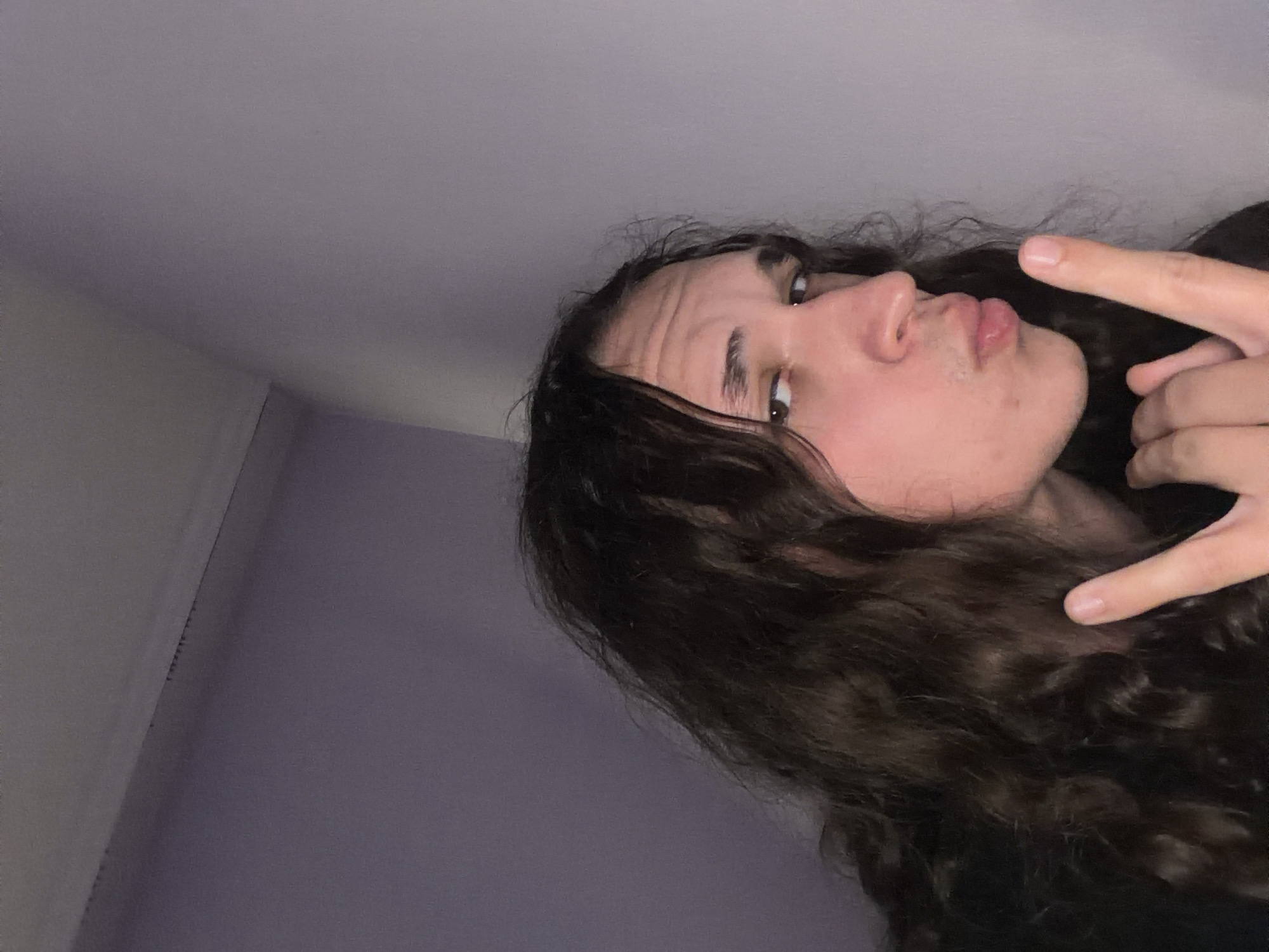 A selfie angled upwards shows a person with long, curly brown hair. The person is looking at the camera with a slight pout and has one hand raised in a "rock on" gesture. The background appears to be an interior, possibly a room, with white and purple walls. There are no indicators of location in Asenovgrad, Bulgaria.