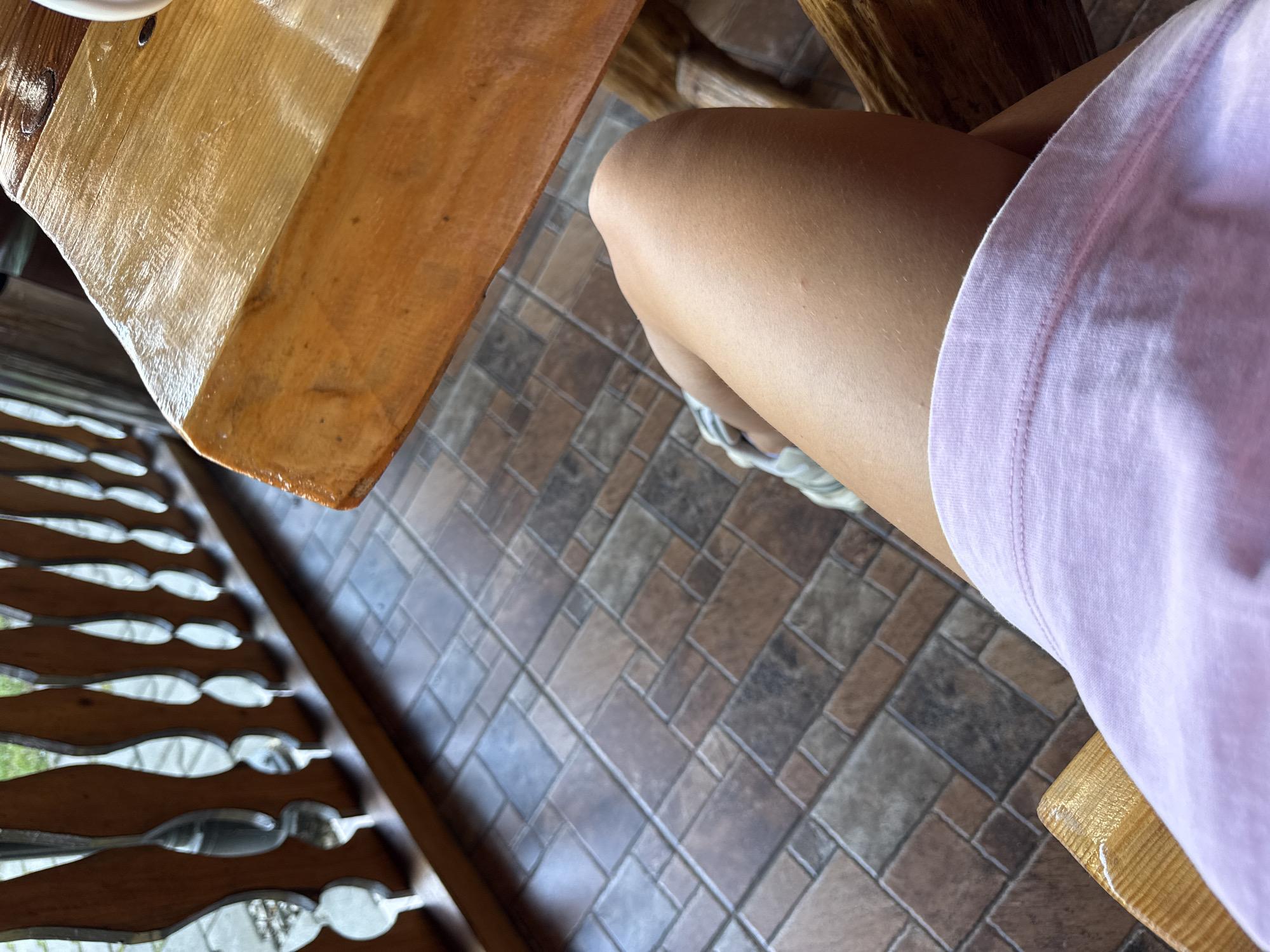 An outdoor tiled patio area is depicted. A person's lap and thighs are visible, clad in a light pink t-shirt. They are seated on a wooden chair. A wooden table is situated to their right, and a wooden railing with decorative spindles runs along the left side of the frame, separating the patio from an area with greenery. The scene is likely located in Cârțișoara, Romania.