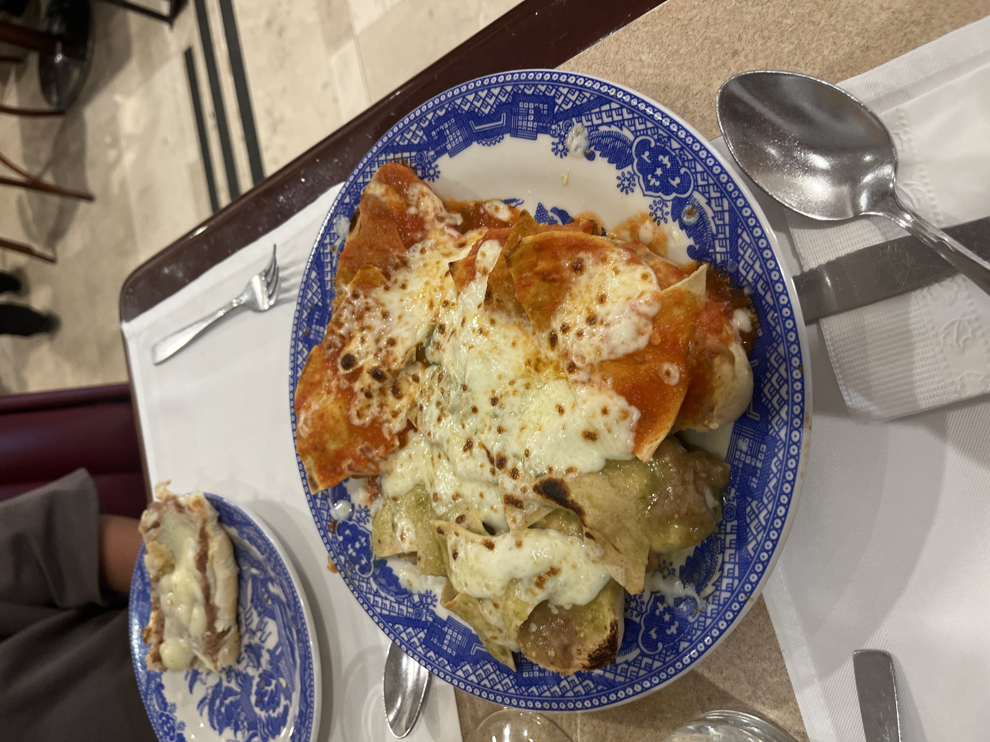 The image shows a plate of enchiladas covered in melted cheese, likely in a restaurant setting. The enchiladas are arranged on a blue and white patterned plate, with some covered in red sauce and others in green sauce. There is a smaller plate with a small portion of enchilada or similar food, also with cheese. Silverware, including a fork and a spoon/knife set, are visible on the white tablecloth surrounding the plates. The location appears to be a restaurant, but the specific city and establishment are unknown. There are chairs in the background, and a brown tray or shelf is partially visible. A person's arm can be seen in the lower left corner.