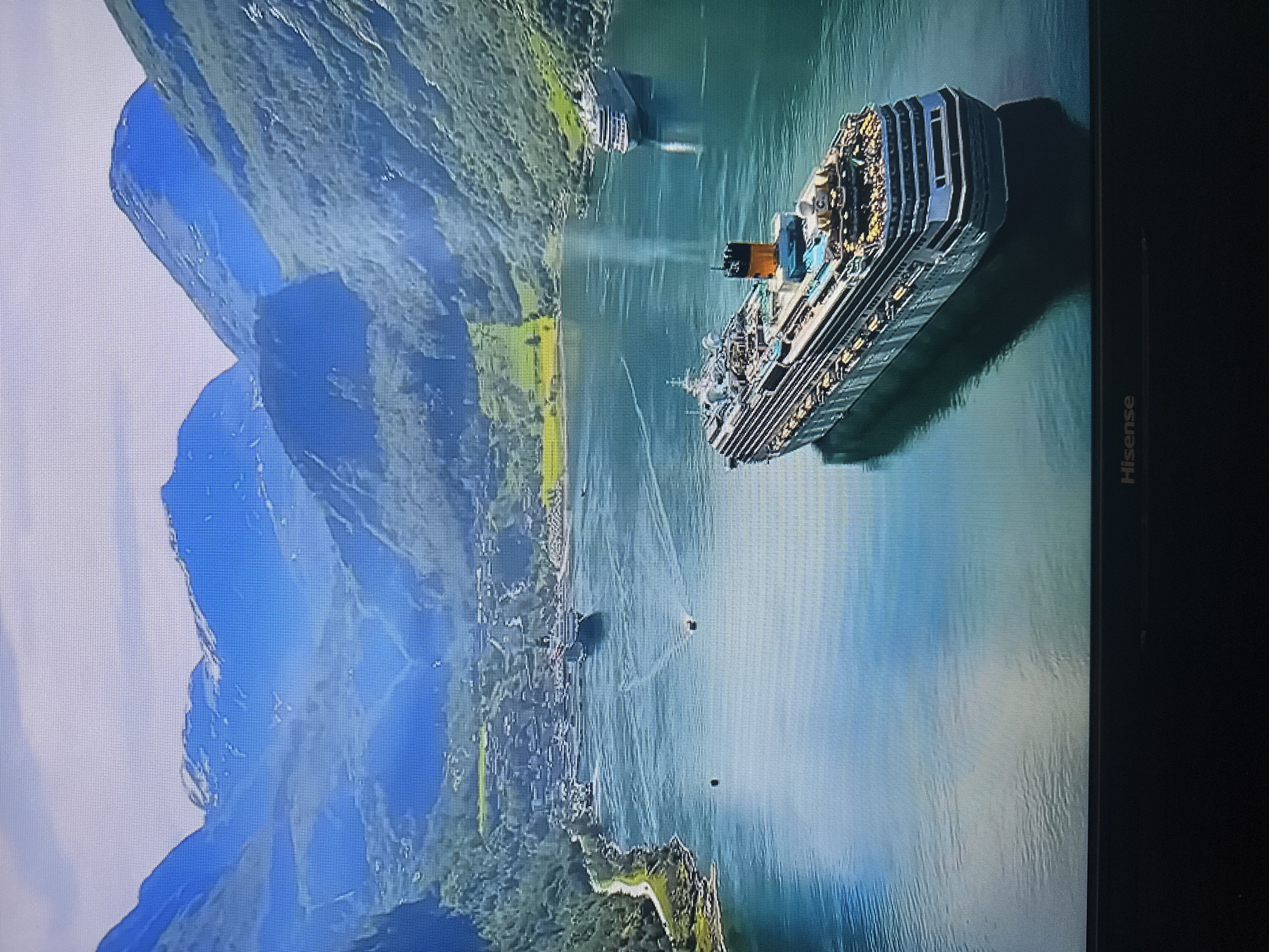The image depicts a panoramic view of a Norwegian fjord with cruise ships navigating the waters. Towering, lush green mountains with patches of snow at their peaks frame a serene, deep blue body of water. A village is nestled along the shoreline, with houses clustered together and a road winding through the landscape. Several cruise ships are visible: one large vessel in the foreground, a second smaller ship further in the distance, and at least one other ship near the village. The water is calm, reflecting the sky and the surrounding mountains. The overall scene is one of natural beauty and human presence within a majestic landscape. The location is not Kyiv, Ukraine.