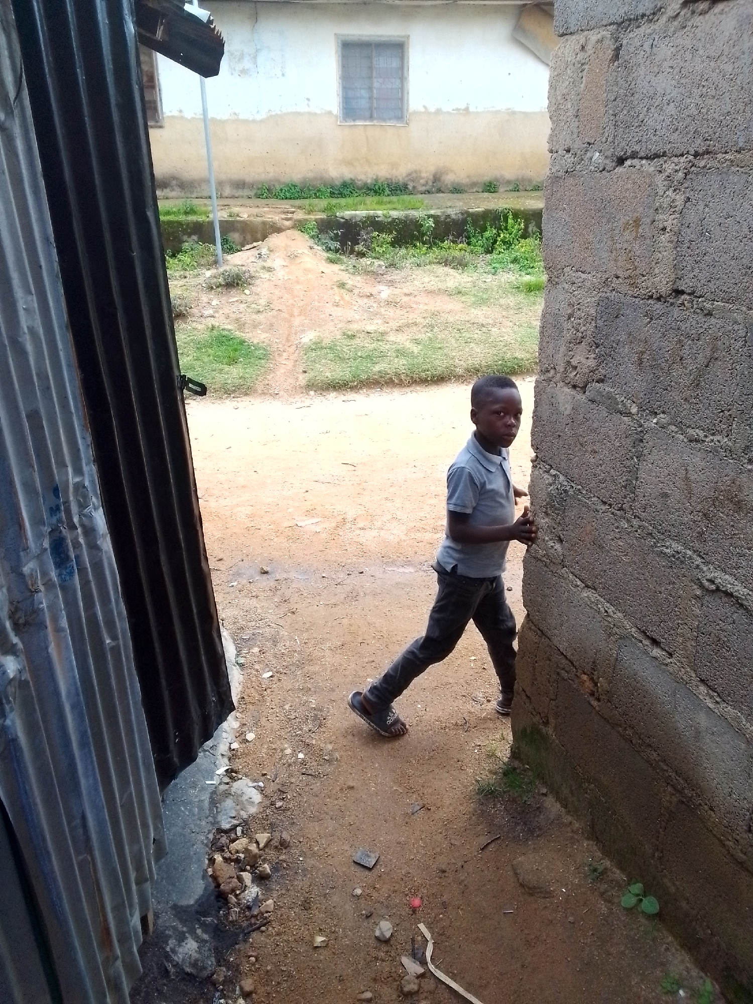 A young Black boy wearing a grey polo shirt and dark pants is shown in a side view, looking towards the camera. He is leaning against a textured concrete block wall, with his right hand on the wall and his left arm extended forward. The boy's feet are visible, one wearing a dark sandal with a white logo. He is standing on a dirt path with some patches of grass. To the left of the boy is a corrugated metal structure with vertical ridges, casting a shadow. In the background, a light-colored building with a window is visible, along with more grass and a dirt path leading away. The scene appears to be outdoors in a somewhat unkempt environment.