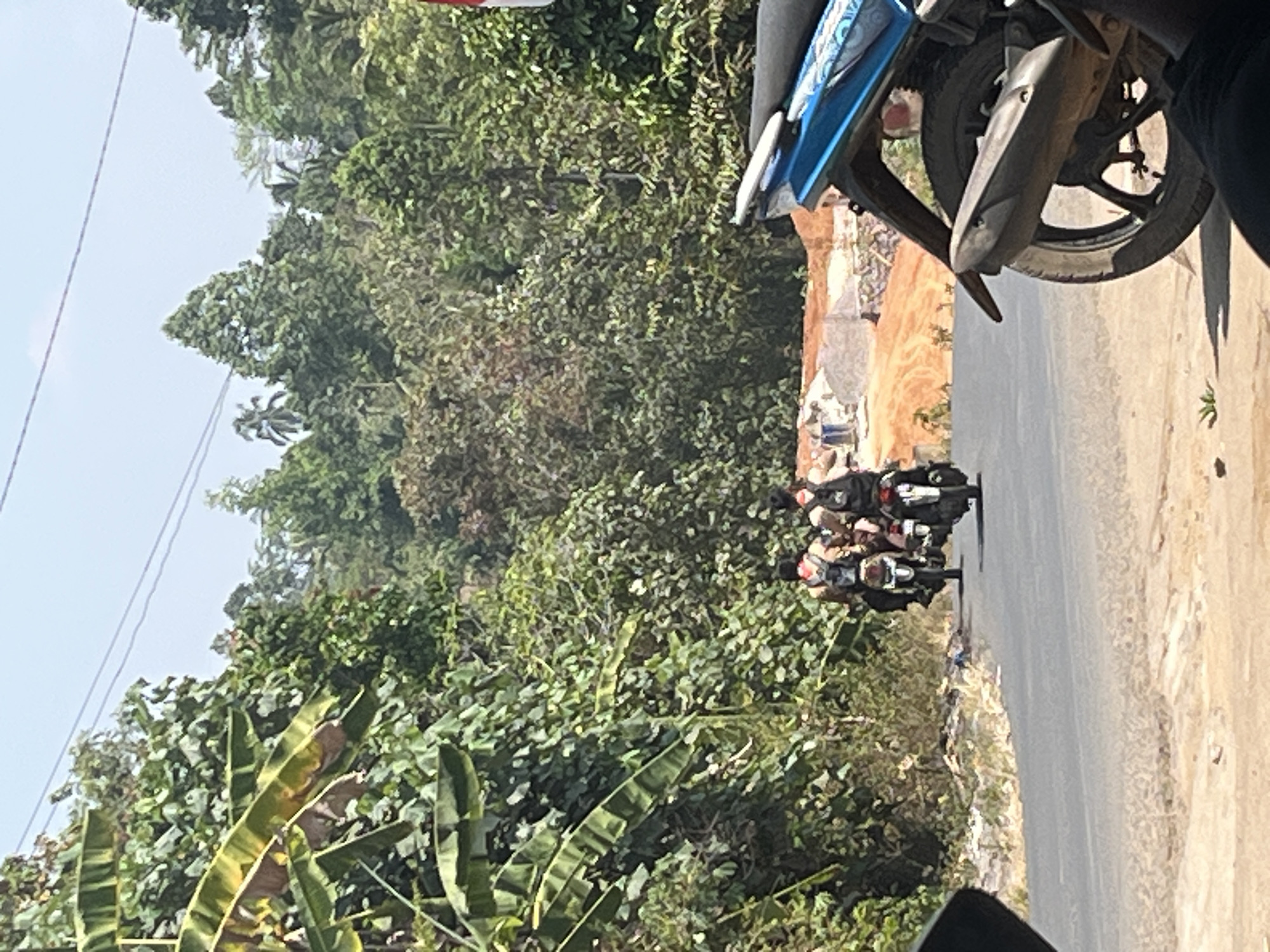 In Karang Sari, Indonesia, a bright, sunny day is depicted on a paved road bordered by dense green foliage. Two individuals are riding motorcycles, one behind the other, heading away from the camera. Both riders are wearing backpacks, and the lead rider has a red bandana around their neck. The road appears somewhat dusty, with a rough, tan-colored shoulder. In the background, a construction site with disturbed orange earth is visible, surrounded by trees and a bright blue sky with power lines. In the foreground, the rear wheel and exhaust of a blue motorcycle are prominently featured. The scene suggests a rural or semi-rural setting outside of a city center.