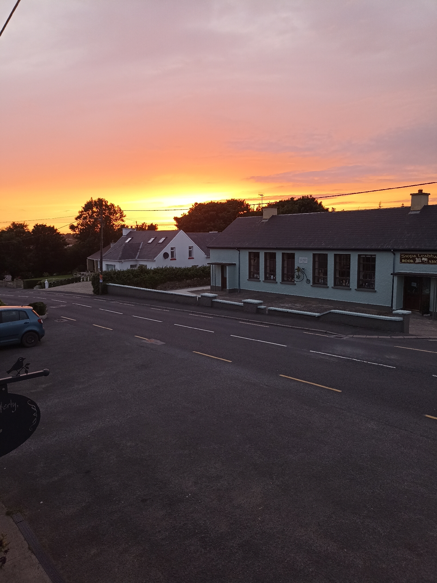 The image captures a tranquil sunset scene on a road in Annagry, Ireland. The sky displays a gradient of colors, transitioning from a soft pink at the top to vibrant orange and yellow hues near the horizon, suggesting the sun is either rising or setting.
In the foreground, a paved road with visible white and yellow lane markings stretches into the distance. A blue car is parked on the left side of the road, and a dark metal sign with a bird silhouette and some text is partially visible in the lower left corner.
On the right side of the road, there are two buildings. The larger building, painted a light blue, features several windows and a sign that reads "SIOPA LEADHBH BOOK SHOP." Adjacent to it, a white house with a dark roof and dormer windows is visible, surrounded by greenery and hedges.
The environment is characterized by trees and vegetation, indicating a rural or semi-rural setting. The overall impression is one of peacefulness and quiet, with the warm glow of the sunset illuminating the scene. There are no people visible in the image, and no specific actions or interactions are depicted. The image is captured from a slightly elevated viewpoint, looking down onto the road and the buildings.