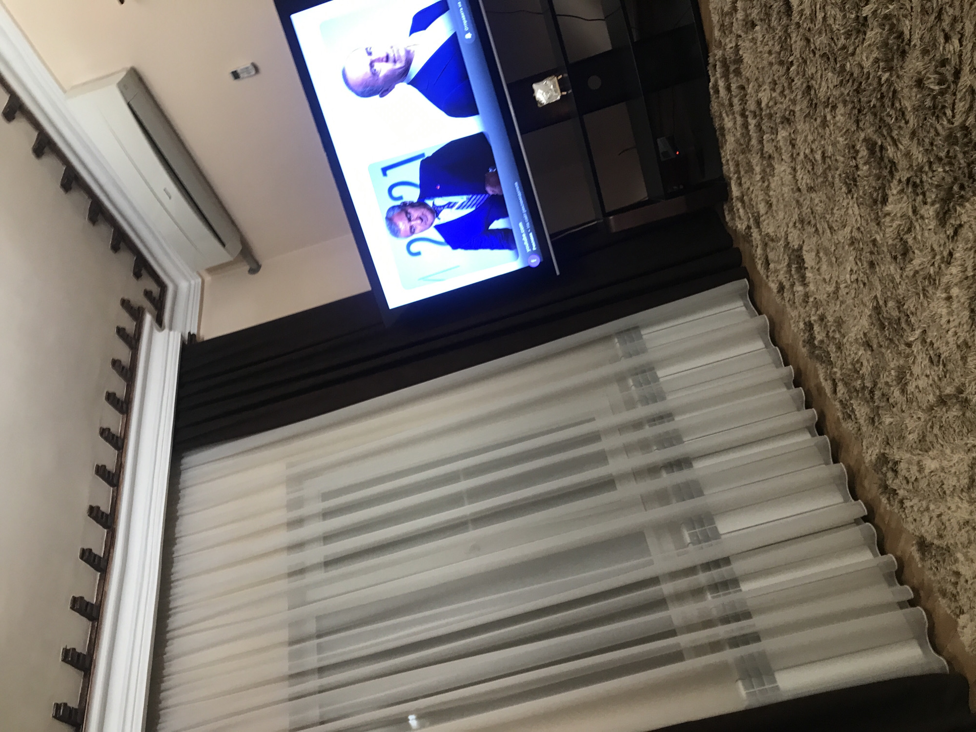 The image shows the interior of a room. A flat-screen television is mounted on the wall, displaying two men in formal attire. Below the television are window blinds. The room is equipped with an air conditioning unit. A carpet is visible on the right side of the image. There is no indication of the location being Yerevan, Armenia.