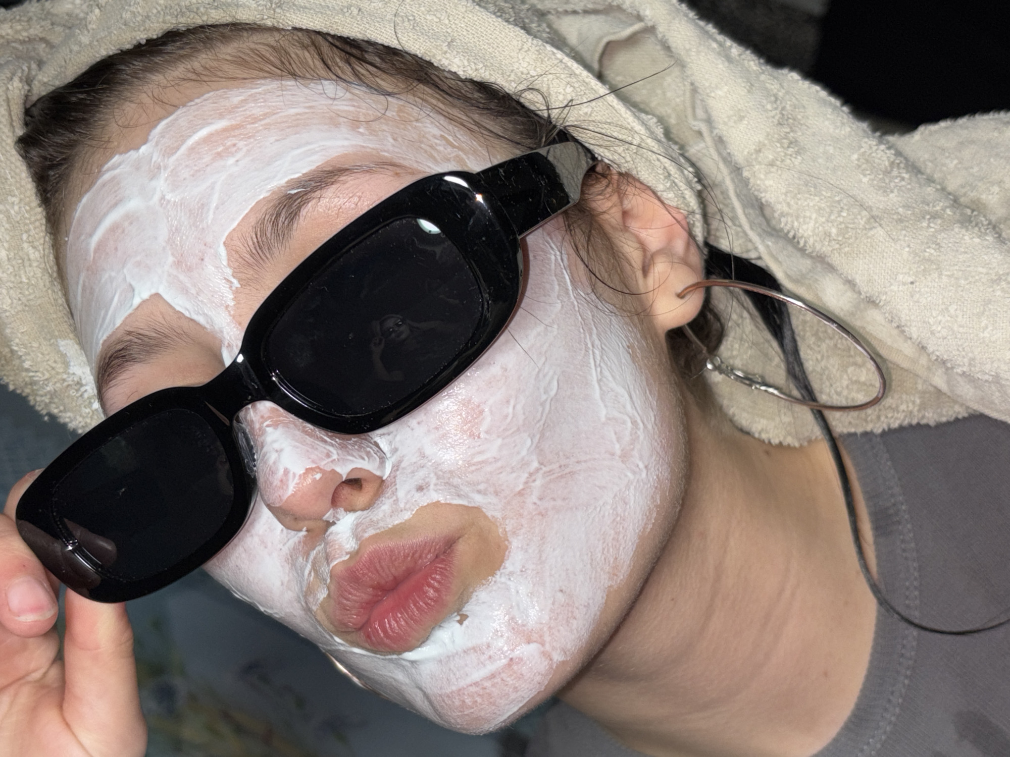 A person is depicted from the shoulders up, face covered in a thick white cream and wearing black sunglasses. The person is wearing a gray shirt and a towel is wrapped around their head. An earring is visible in the left ear. The location cannot be determined from the image. The city of Фоки, Russia, cannot be verified as the location.