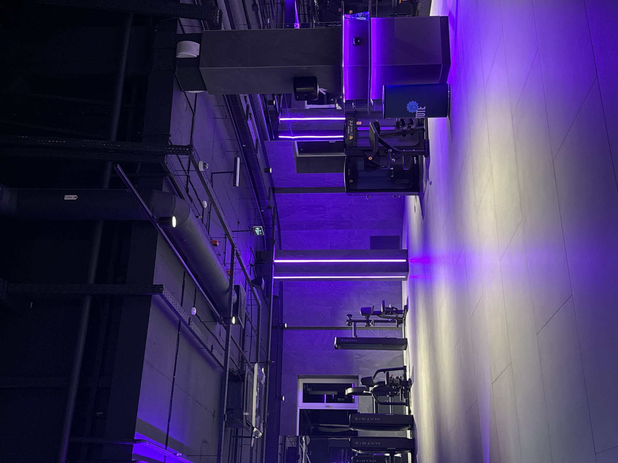 The image depicts a modern, well-lit gymnasium interior with purple ambient lighting. The floor is tiled with large, light-colored rectangular tiles. The walls have a textured, grey finish. Various pieces of gym equipment, including weight machines and cardio equipment, are visible, some bearing the "MATRIX" logo. A large rectangular pillar with integrated lighting and shelving is prominent on the right side of the frame. A green cylindrical trash bin with the word "TOTE" printed on it is situated near the pillar. The ceiling is industrial in design, with exposed pipes, ventilation systems, and lighting fixtures. There are no people visible in the image. The scene suggests a contemporary fitness facility. The image does not provide any explicit information about its specific location in Krasnoyarsk, Russia, nor does it offer any context to infer such a detail.