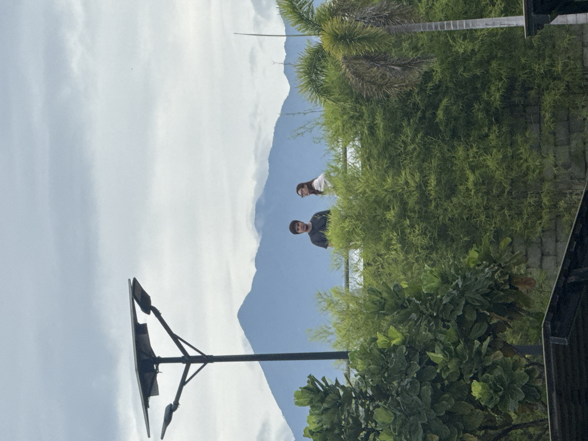 Two people are on a rooftop in Davao City, Philippines, with a mountain in the background. The scene includes lush green foliage, a palm tree, and a streetlight. The sky is cloudy.